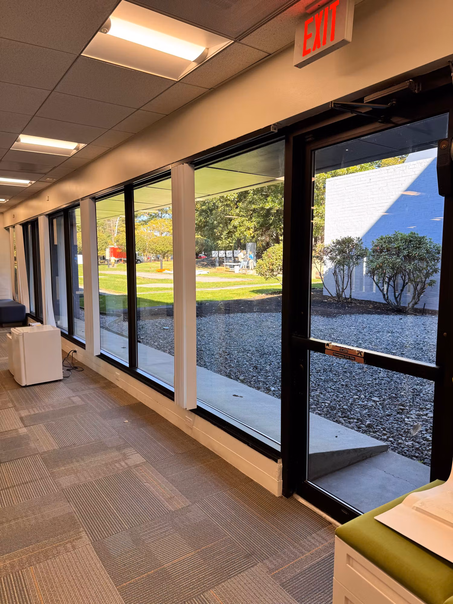 Office corridor with floor-to-ceiling tinted windows and a glass exit door looking out to a lawn.