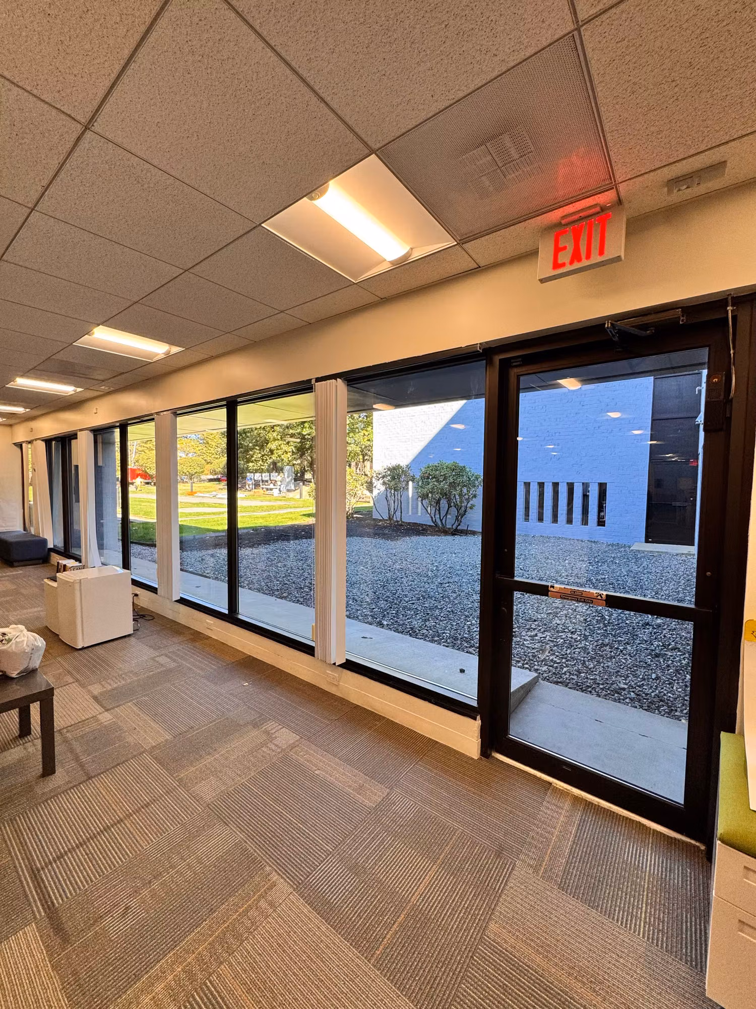 Office lounge corridor lined with large tinted windows and a glass exit door overlooking the grounds.