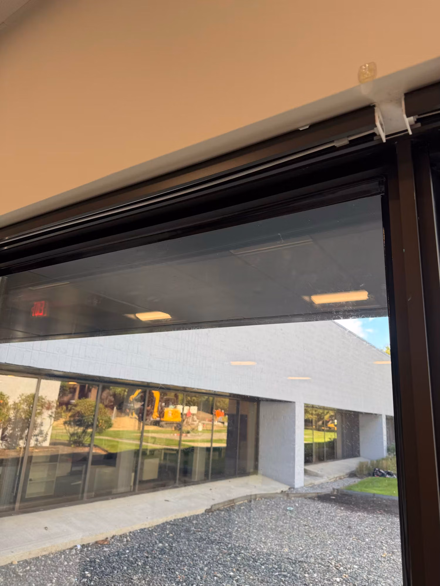 Floor-to-ceiling office window with a dark solar screen cutting glare, overlooking a brick building and green grounds.