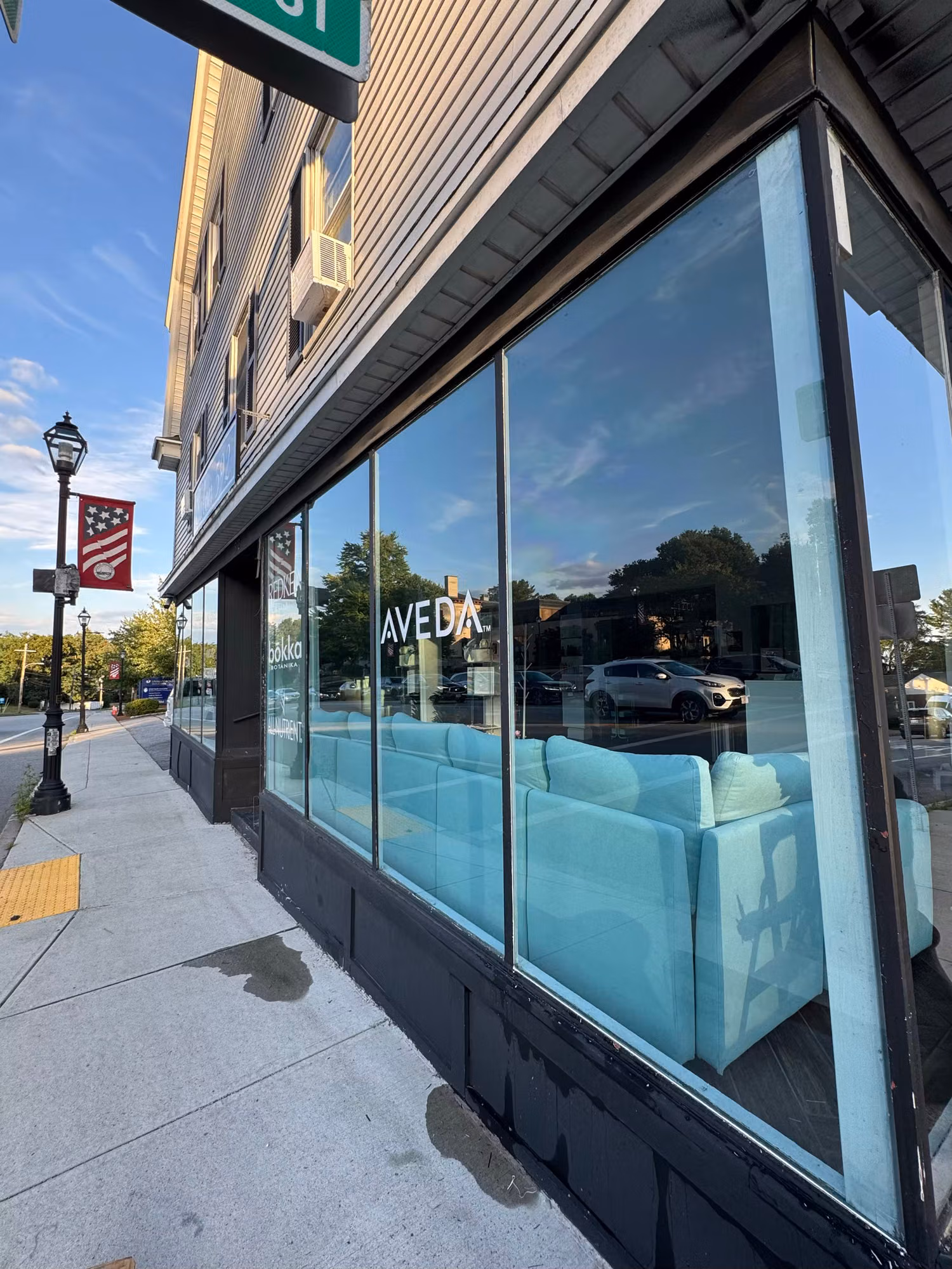 Corner salon storefront with large tinted commercial windows reflecting trees, sky, and passing traffic.
