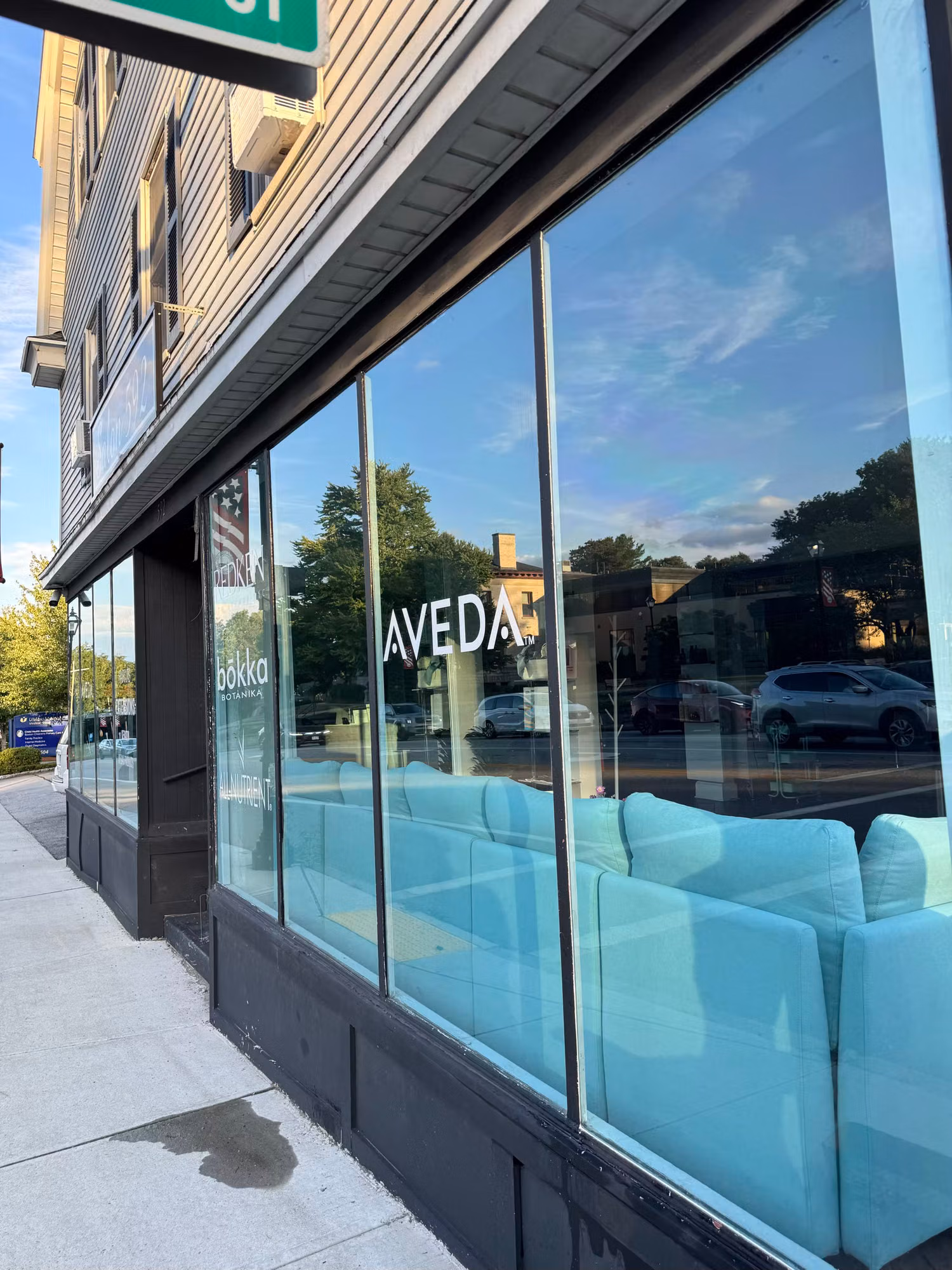 Street-level salon storefront window wall with reflective tinted glass mirroring parked cars and trees outside.