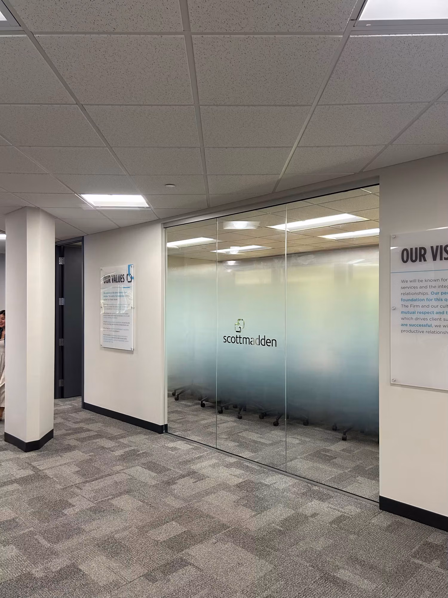 Office corridor glass partition wall with a frosted privacy band and cut-vinyl logo lettering screening the meeting room beyond.