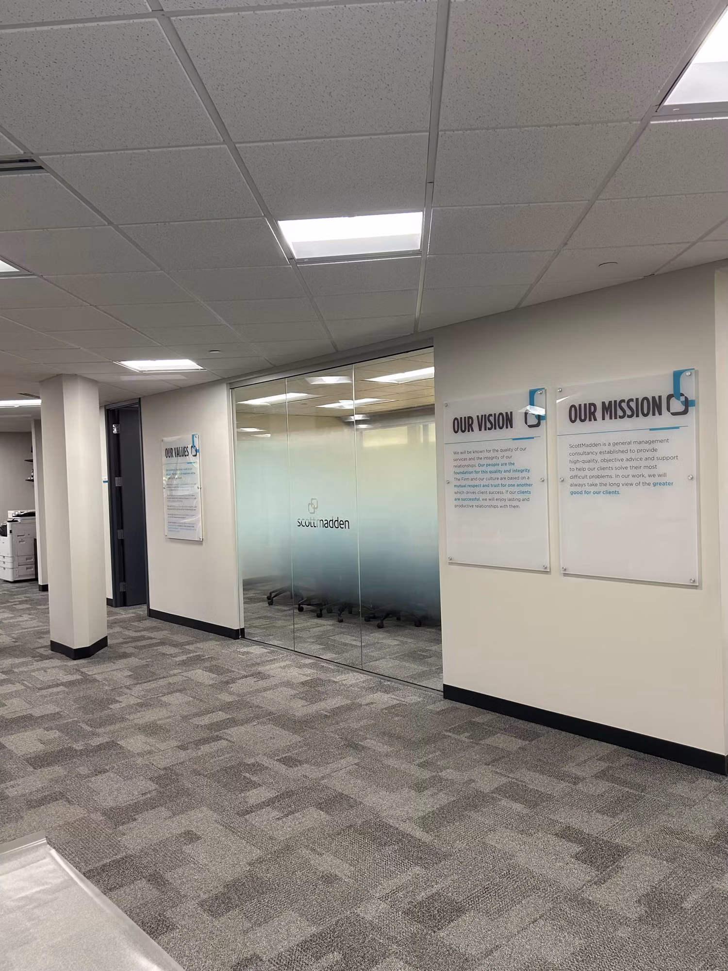 Office corridor glass partition wall with a frosted privacy band and dimensional vision and mission wall graphics.