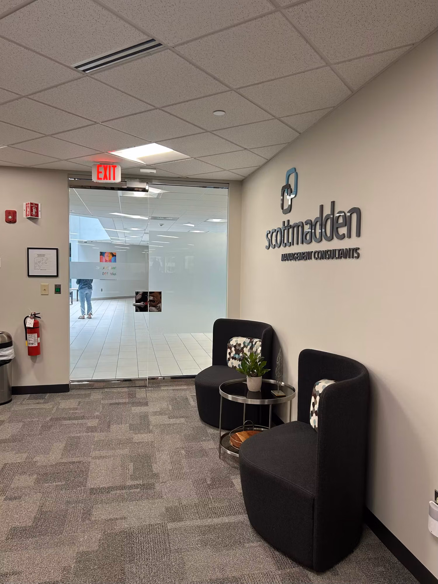 Office reception lobby with a clear glass partition wall and entry door opening into a bright interior workspace.