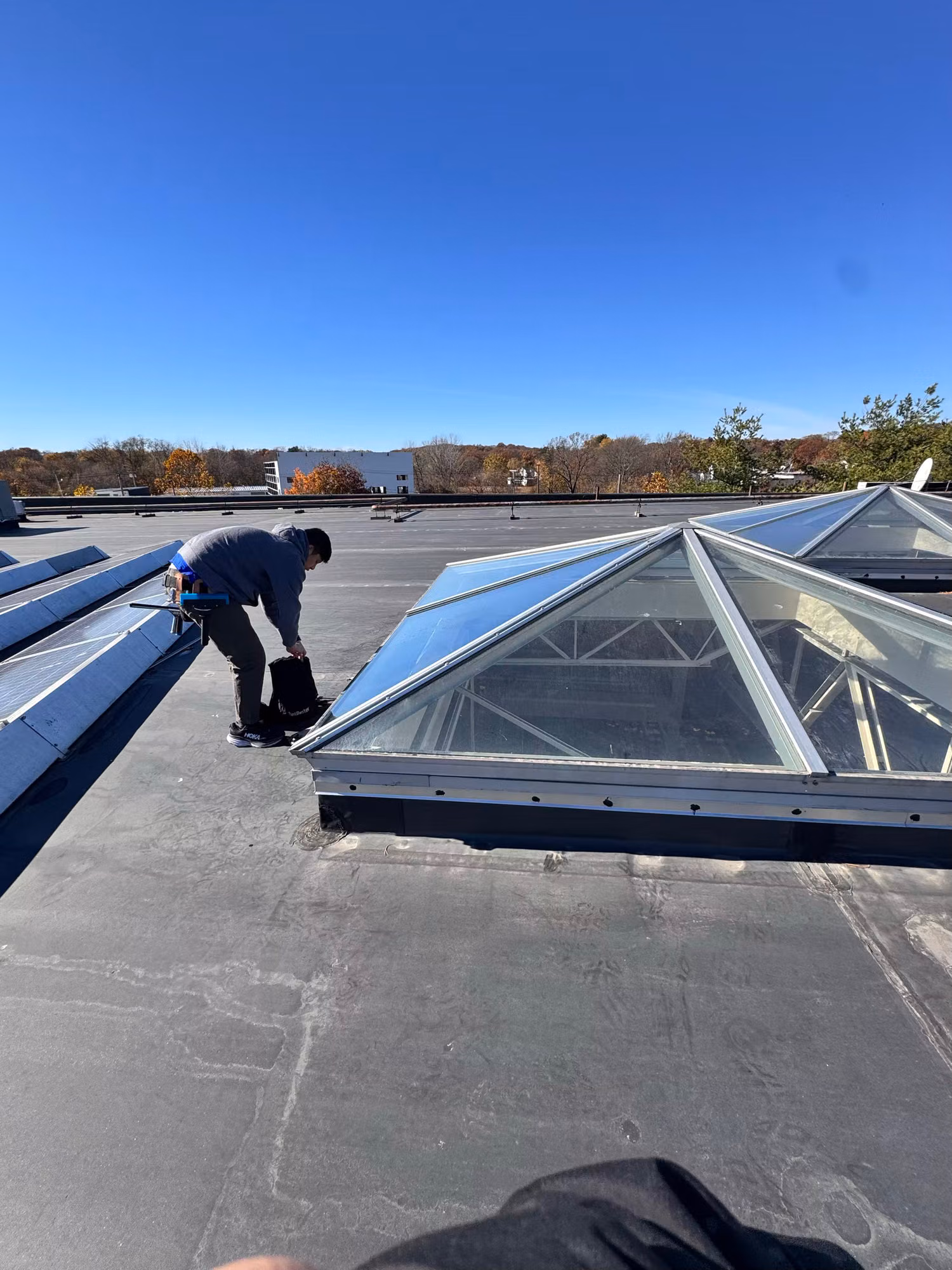 Technician kneeling beside a pyramid glass skylight on a commercial roof while applying window film.