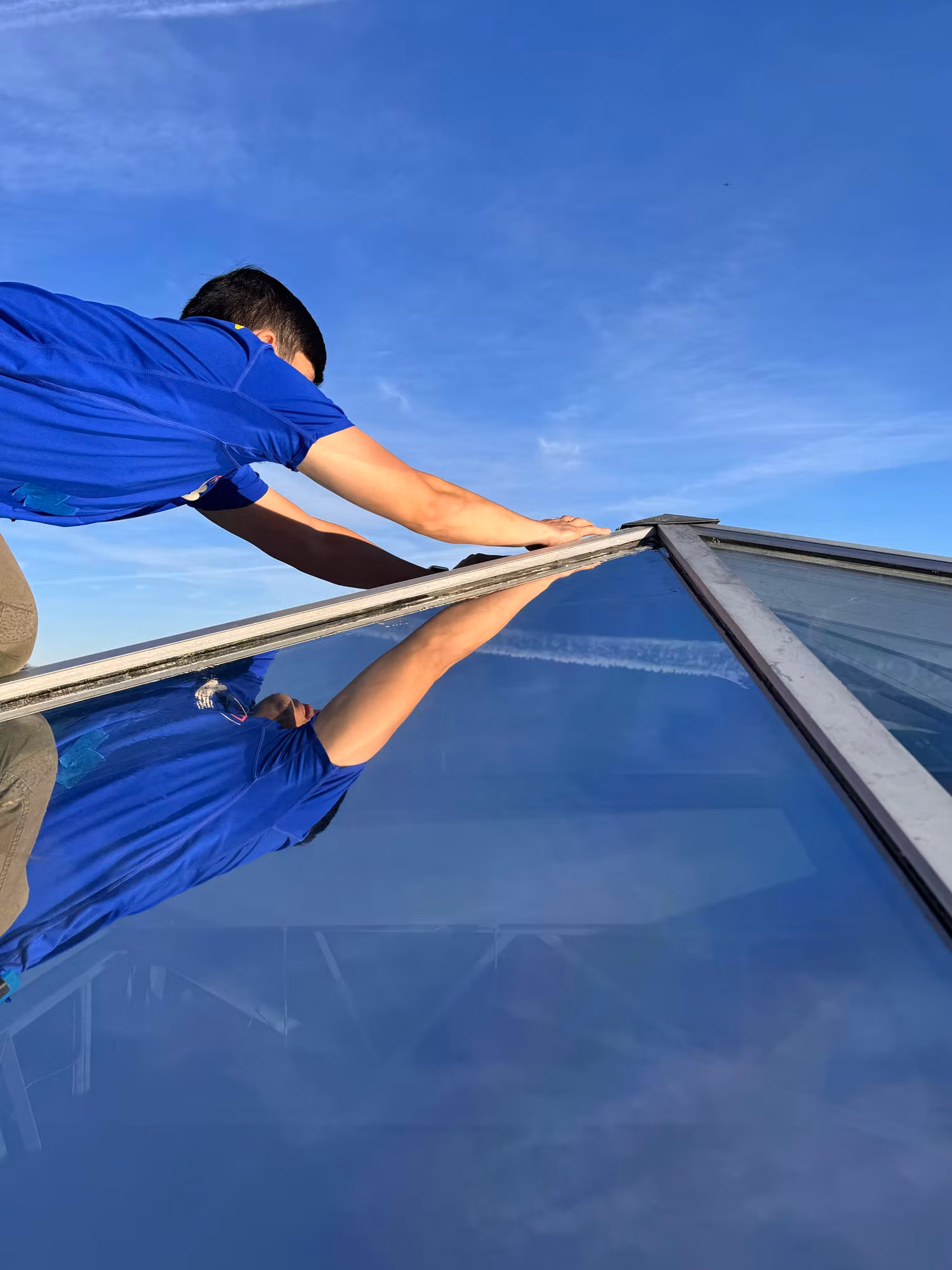 Installer reaching across a sloped glass skylight to smooth window film against a clear blue sky.
