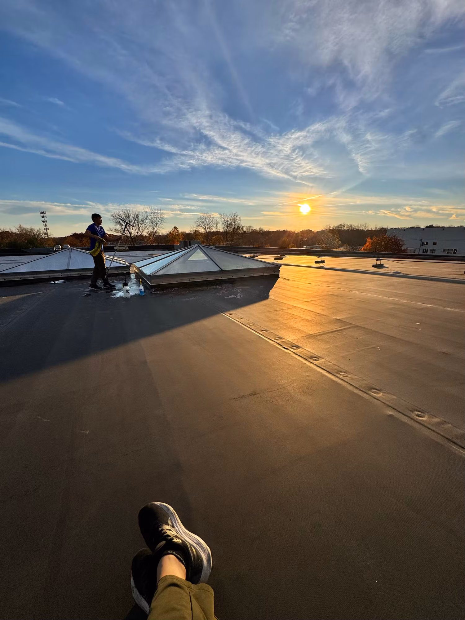Worker tending glass skylights on a flat commercial rooftop as the sun sets in the background.