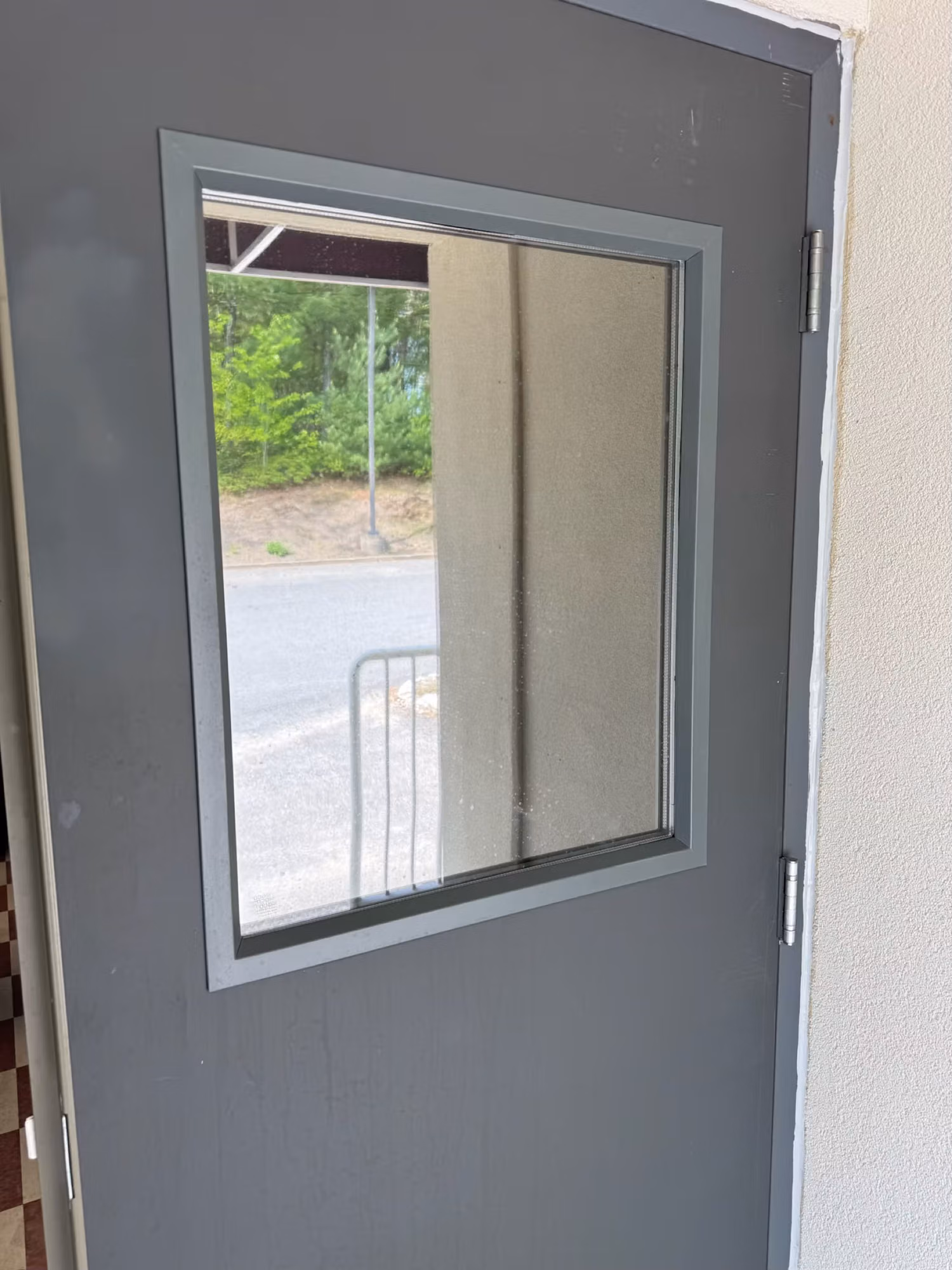 Square glass vision panel on a gray metal door finished with window film, looking out toward trees and pavement.