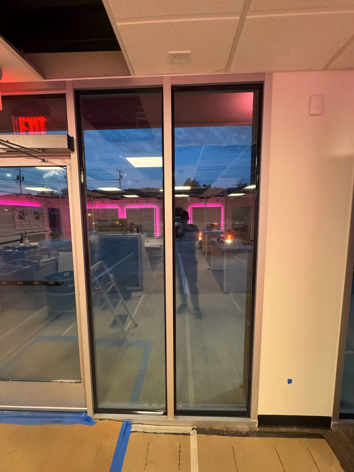 Interior view of tall storefront glass entry doors with an exit sign overhead and painter's tape on the floor during install.