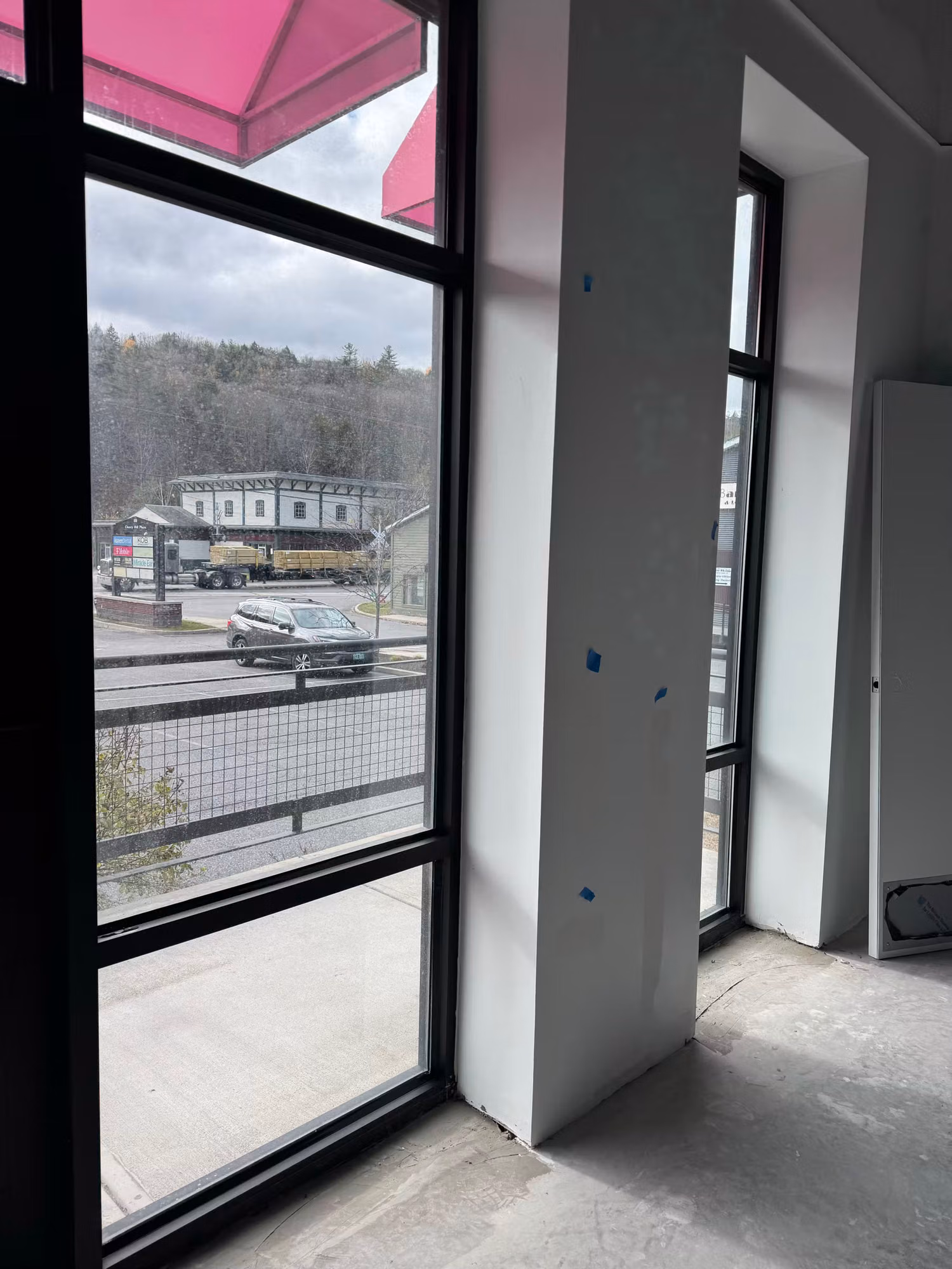 Interior corner of a commercial space under buildout with full-height storefront windows overlooking a roadway.