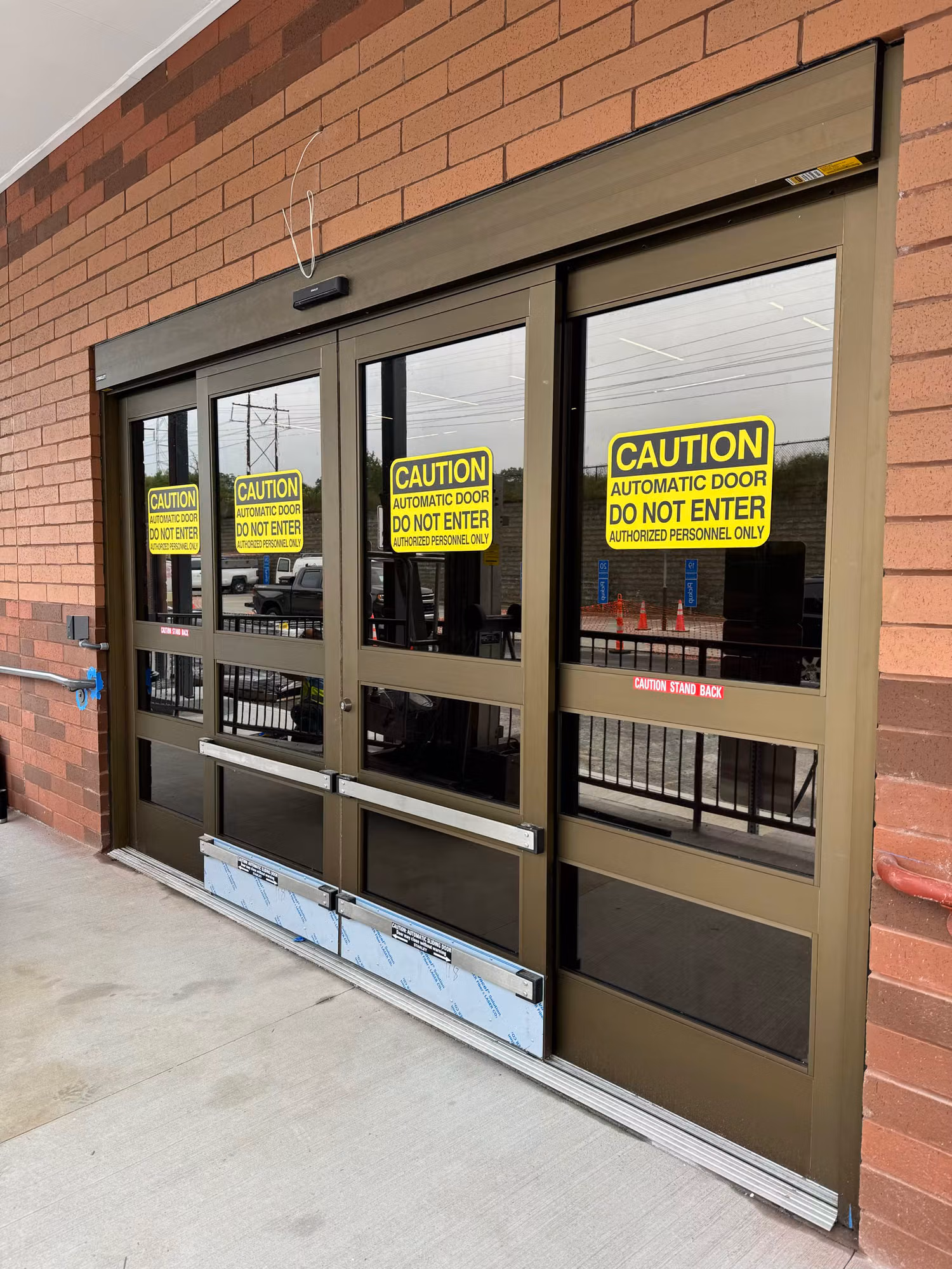 Bronze automatic sliding entry doors with tinted glass and yellow caution signage along a brick storefront wall.