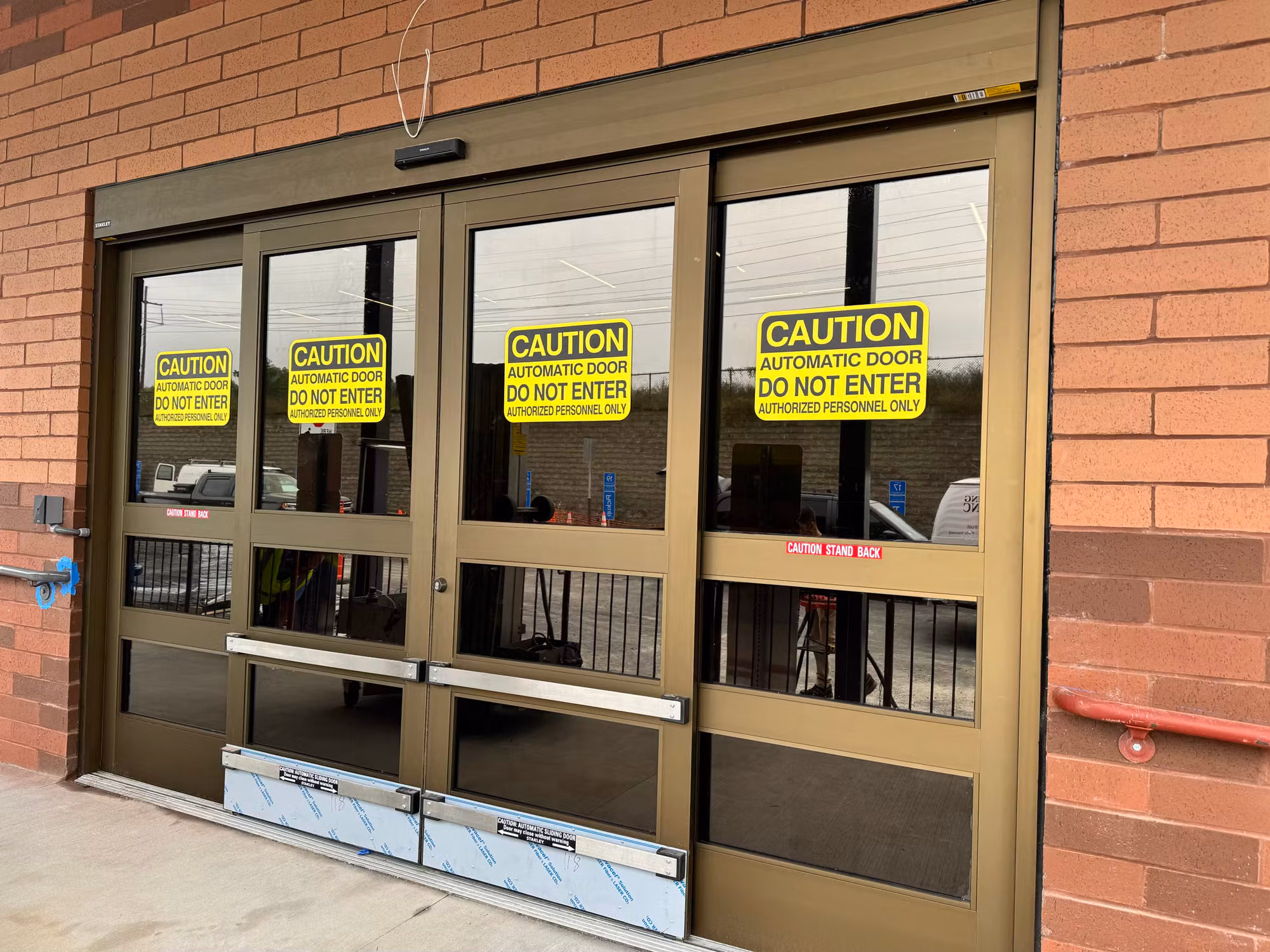 Tinted glass automatic sliding entry doors with yellow caution decals framed in bronze against a brick exterior.