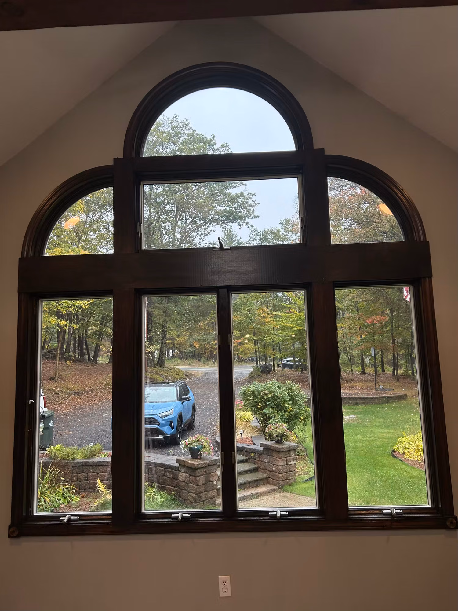 Interior view of a large arched Palladian window with a dark wood frame looking out to trees and a driveway.