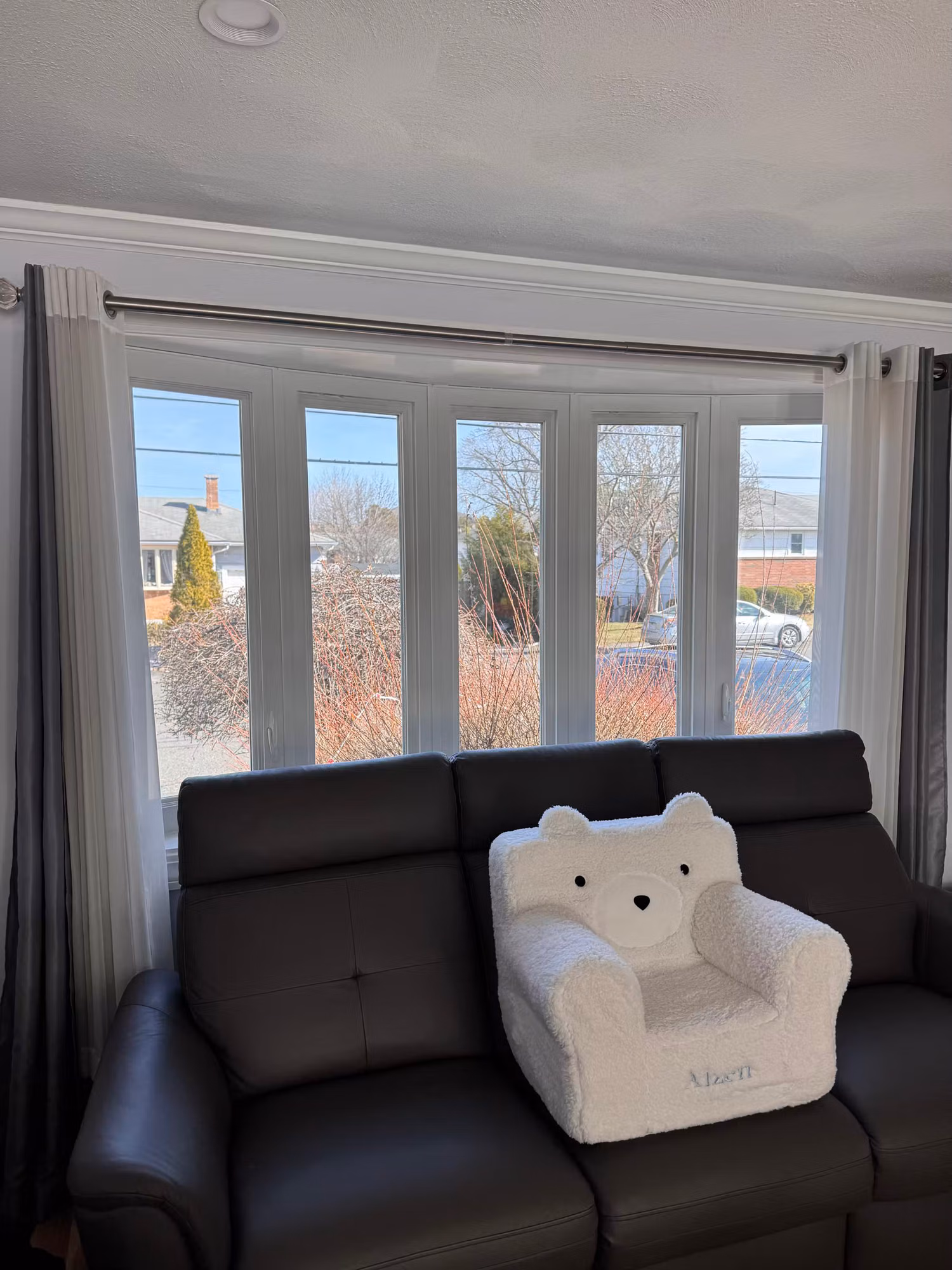 Living room bay window with five angled glass panes overlooking a residential street above a leather sofa.
