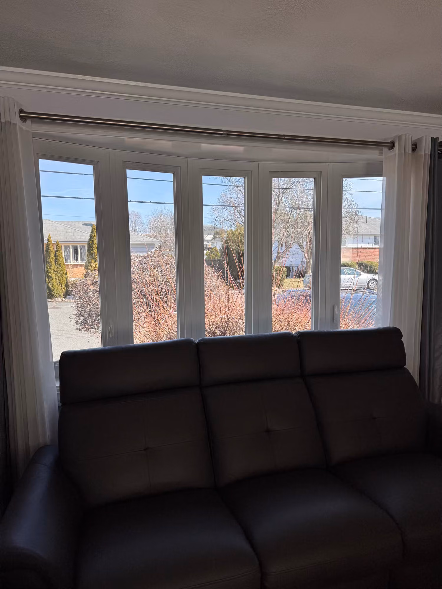 Multi-pane bay window in a living room behind a dark leather sofa with a neighborhood view outside.