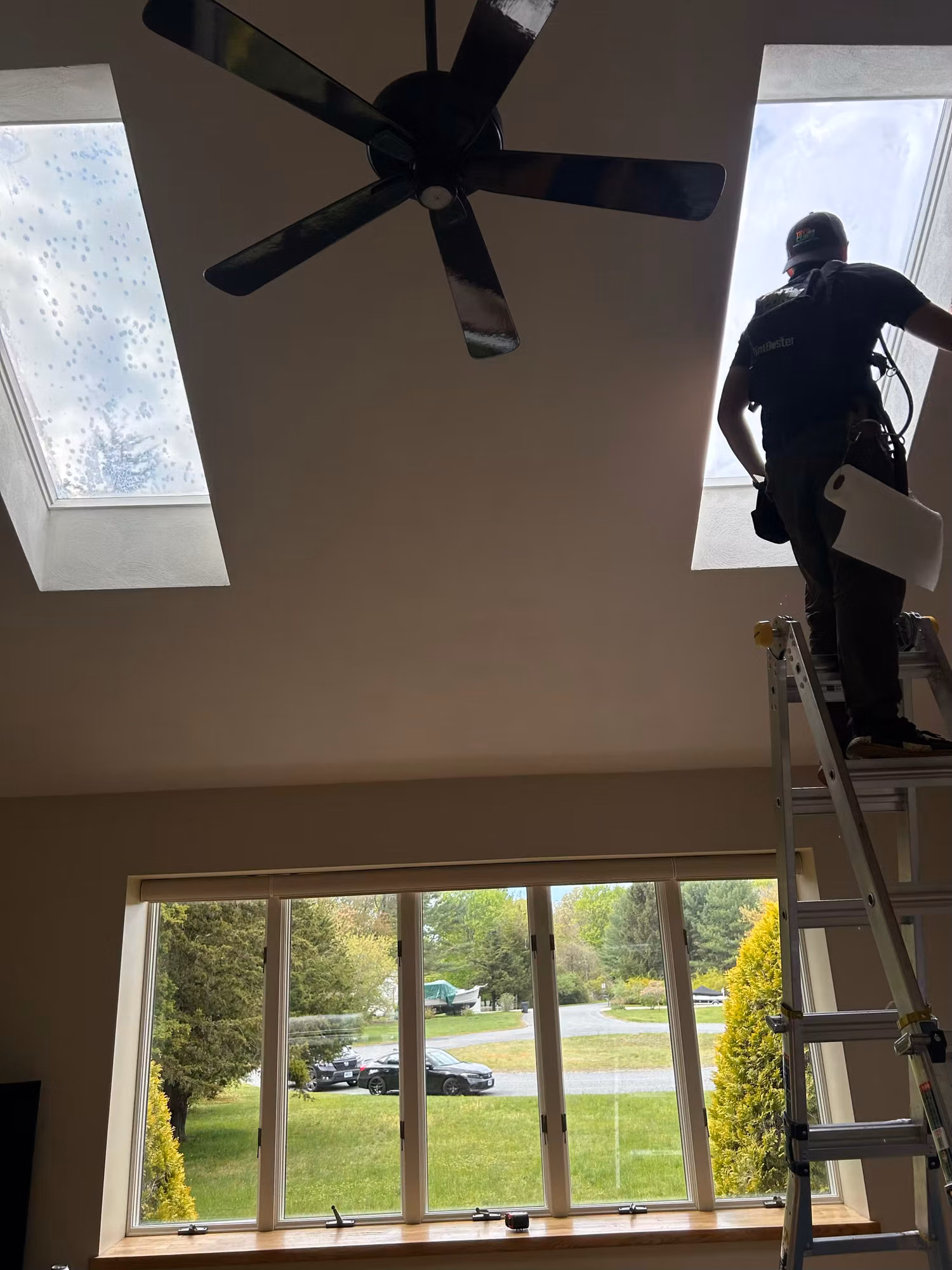 Installer on a ladder applying window film to two vaulted-ceiling skylights above a living room window.