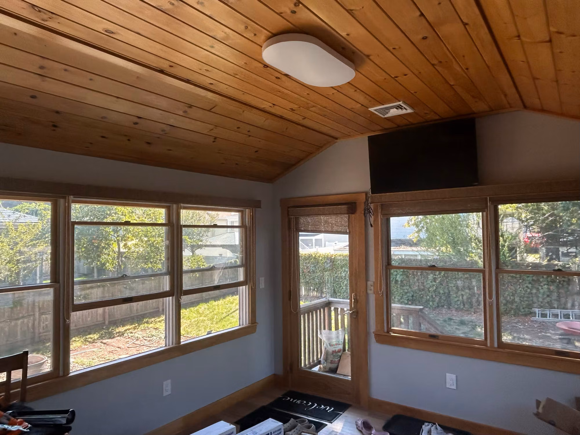 A wraparound bank of sunroom windows under a wood plank ceiling, looking out onto a deck and backyard.