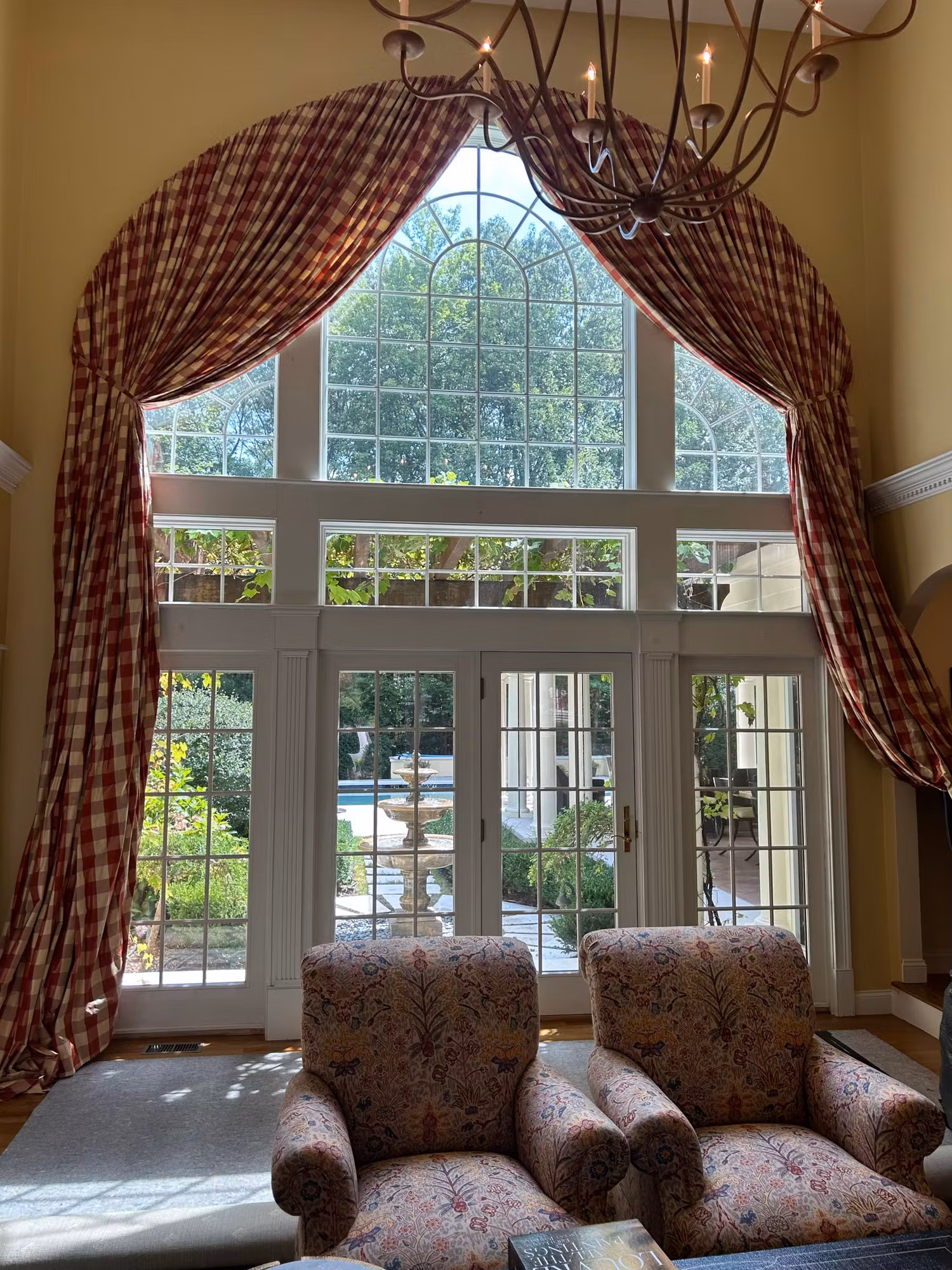 Tall arched Palladian window above French doors in a living room with garden views and gingham drapes.