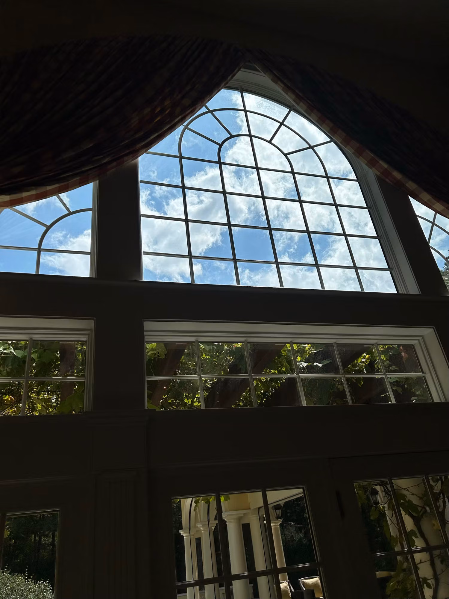 Upper arched transom window with divided grilles letting in blue sky and greenery in a residential room.