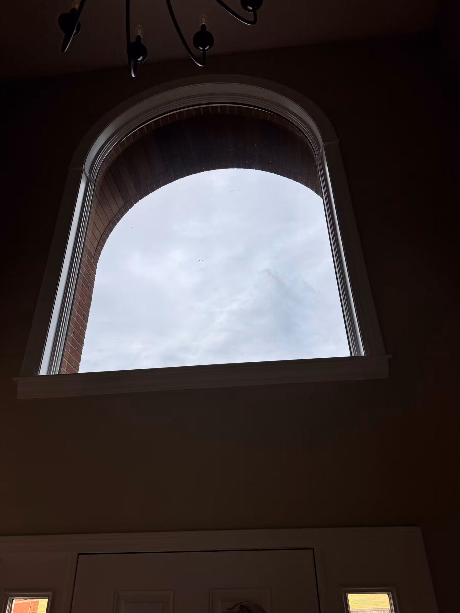 Tall arched foyer window seen from inside beneath a chandelier, clear glass framing an overcast sky.