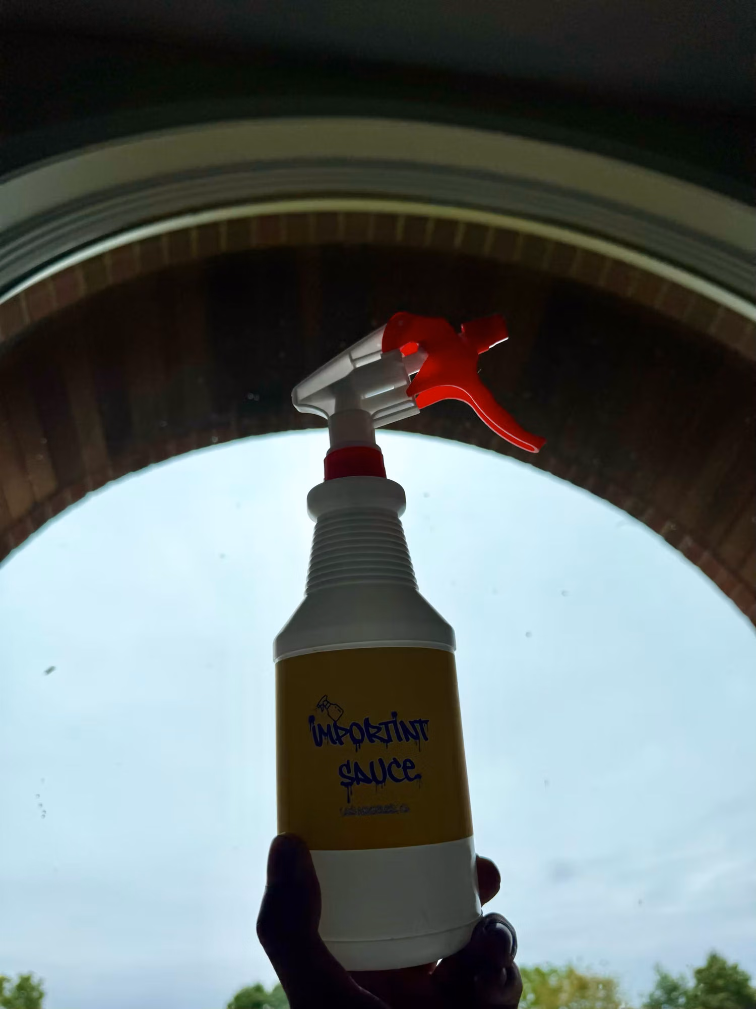 Hand holding a labeled spray bottle of installation solution in front of an arched residential window during a film install.