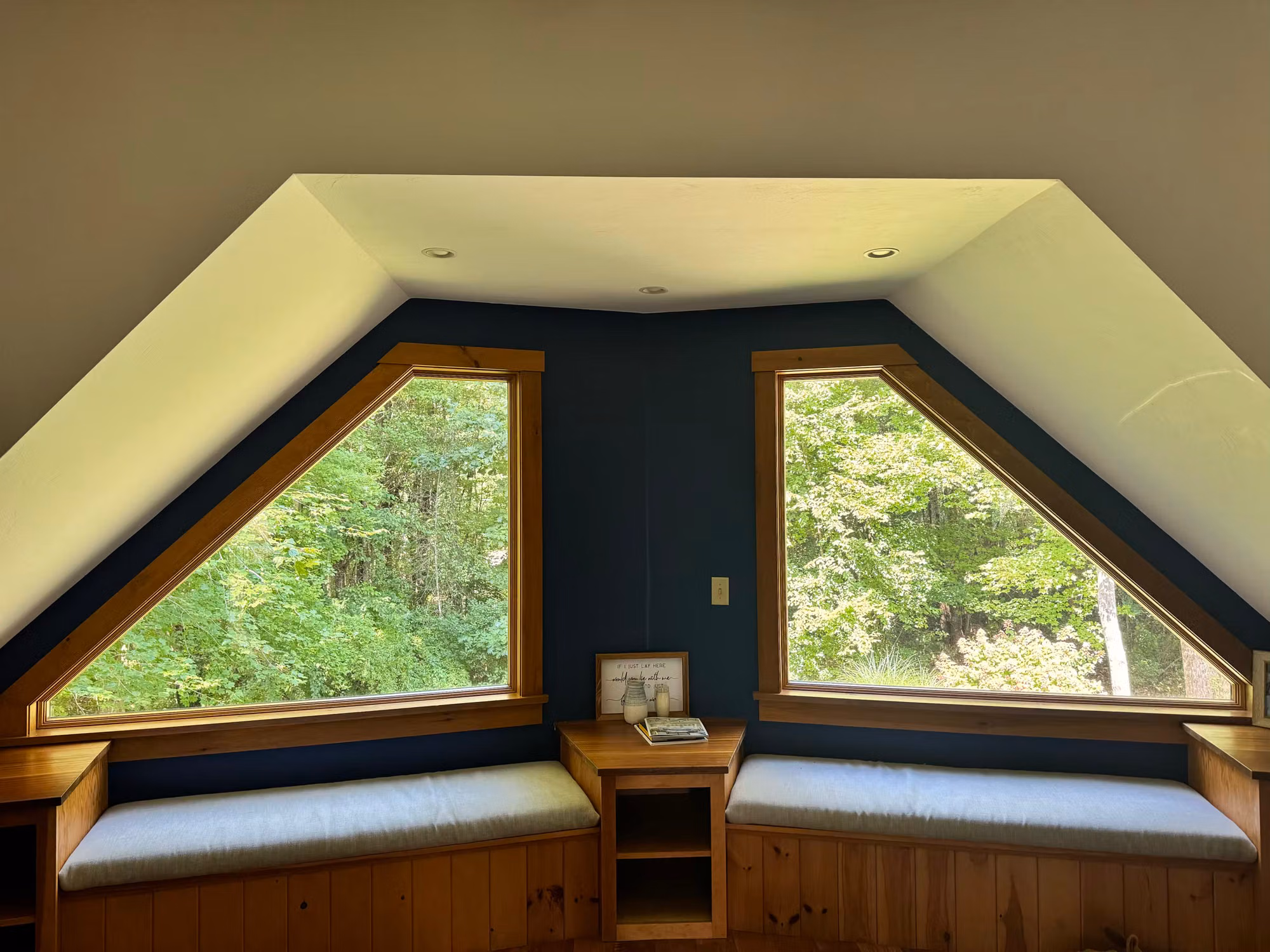 Two triangular dormer windows flanking a built-in window seat in an attic bedroom with navy walls and wood trim.