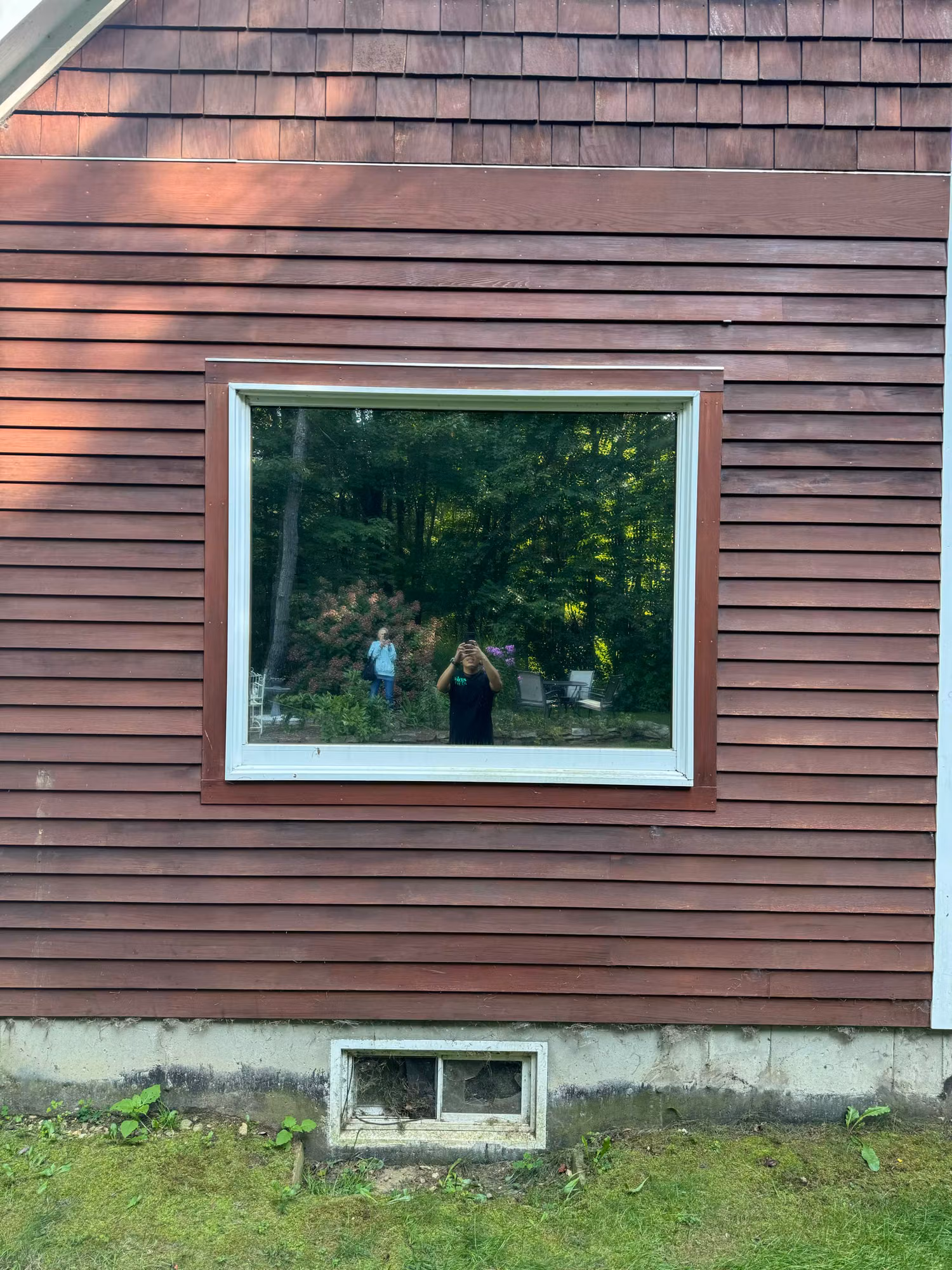 Large picture window on red clapboard siding with a reflective glass surface and surrounding greenery.