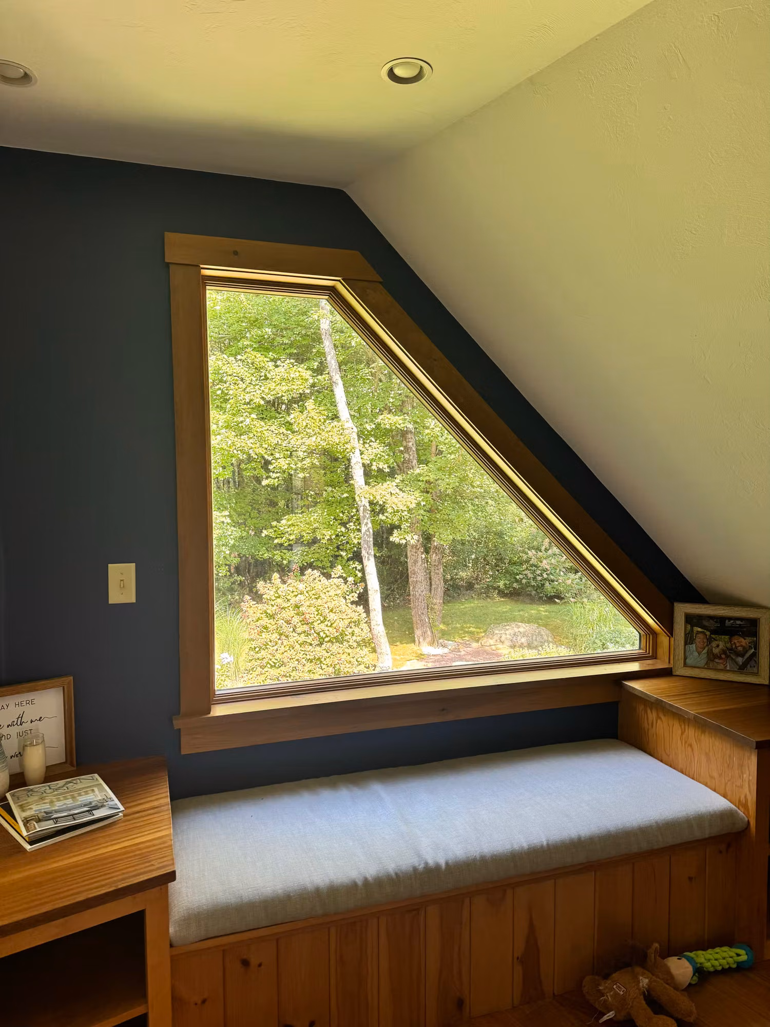 Angled view of a triangular wood-framed window over a cushioned window seat overlooking trees in a loft bedroom.