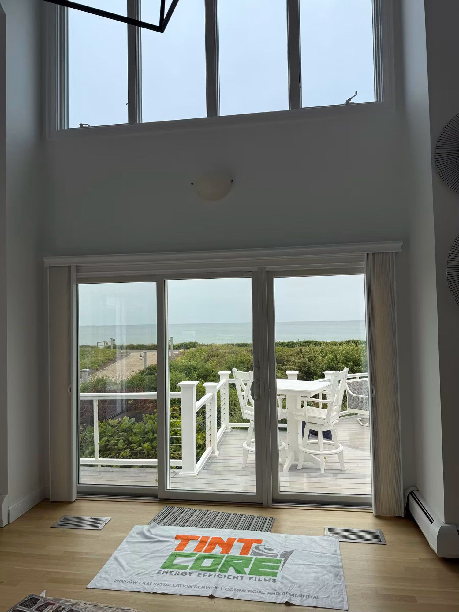 Two-story living room with tall windows and sliding doors to an ocean-view deck during window film installation.