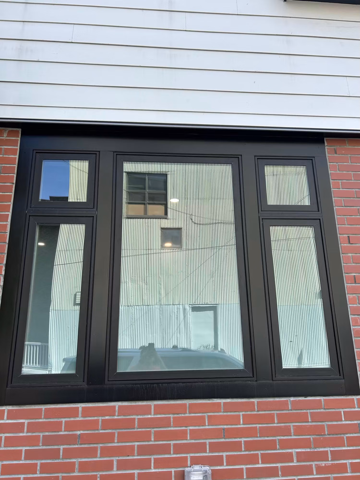 Large black-framed multi-pane picture window on a brick and clapboard home with clear glass reflecting the surroundings.