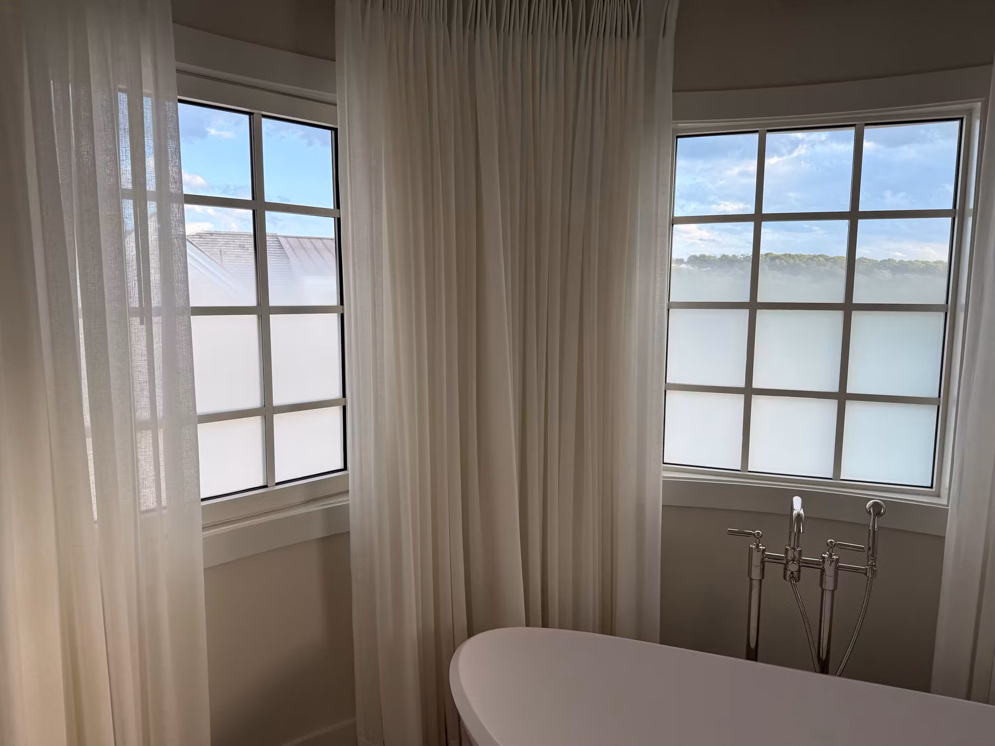 Two divided-light bathroom windows behind sheer curtains with frosted privacy film on the lower panes beside a soaking tub.