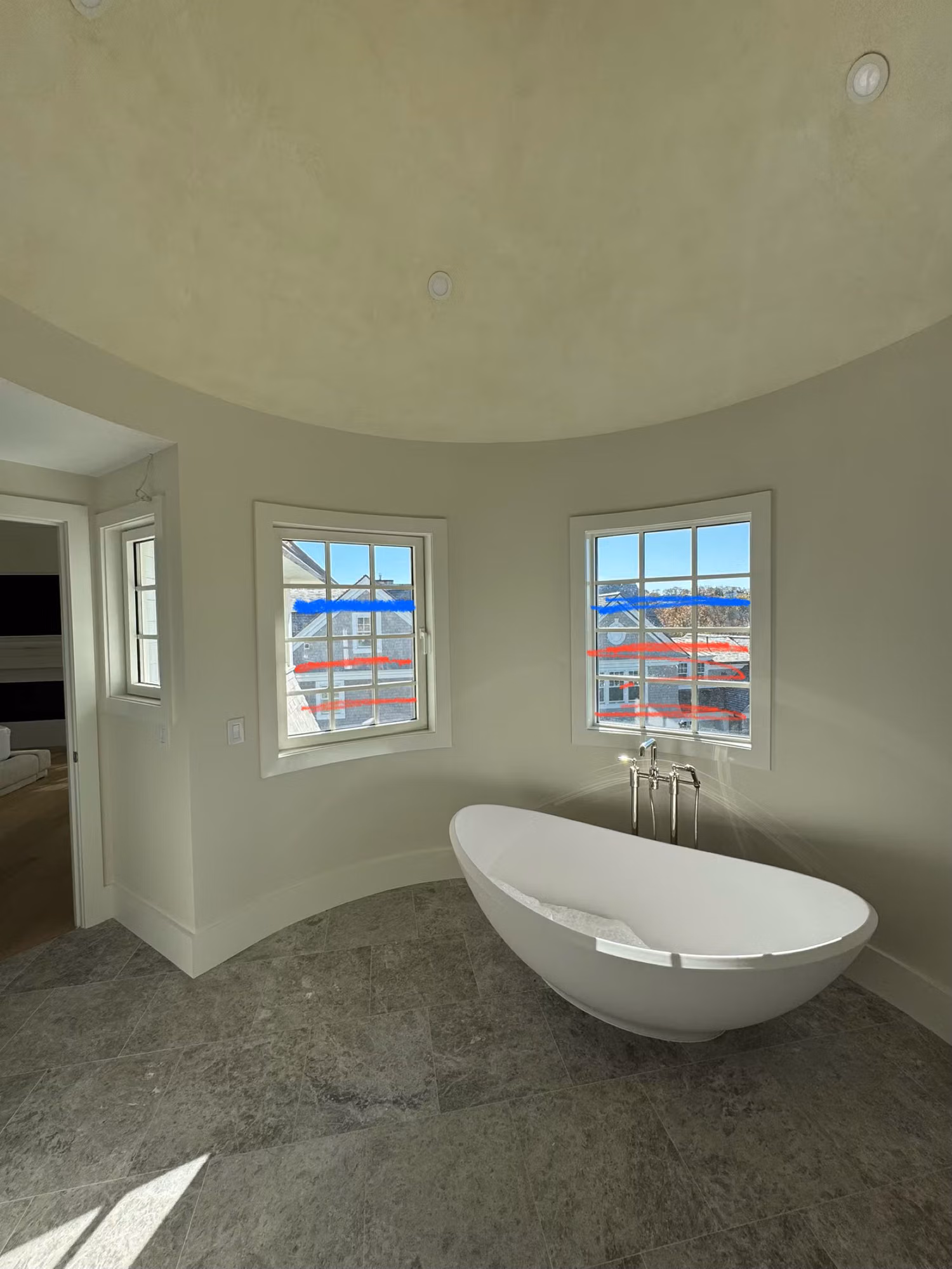 Curved bathroom with a freestanding tub and two divided-light windows looking toward neighboring rooftops.