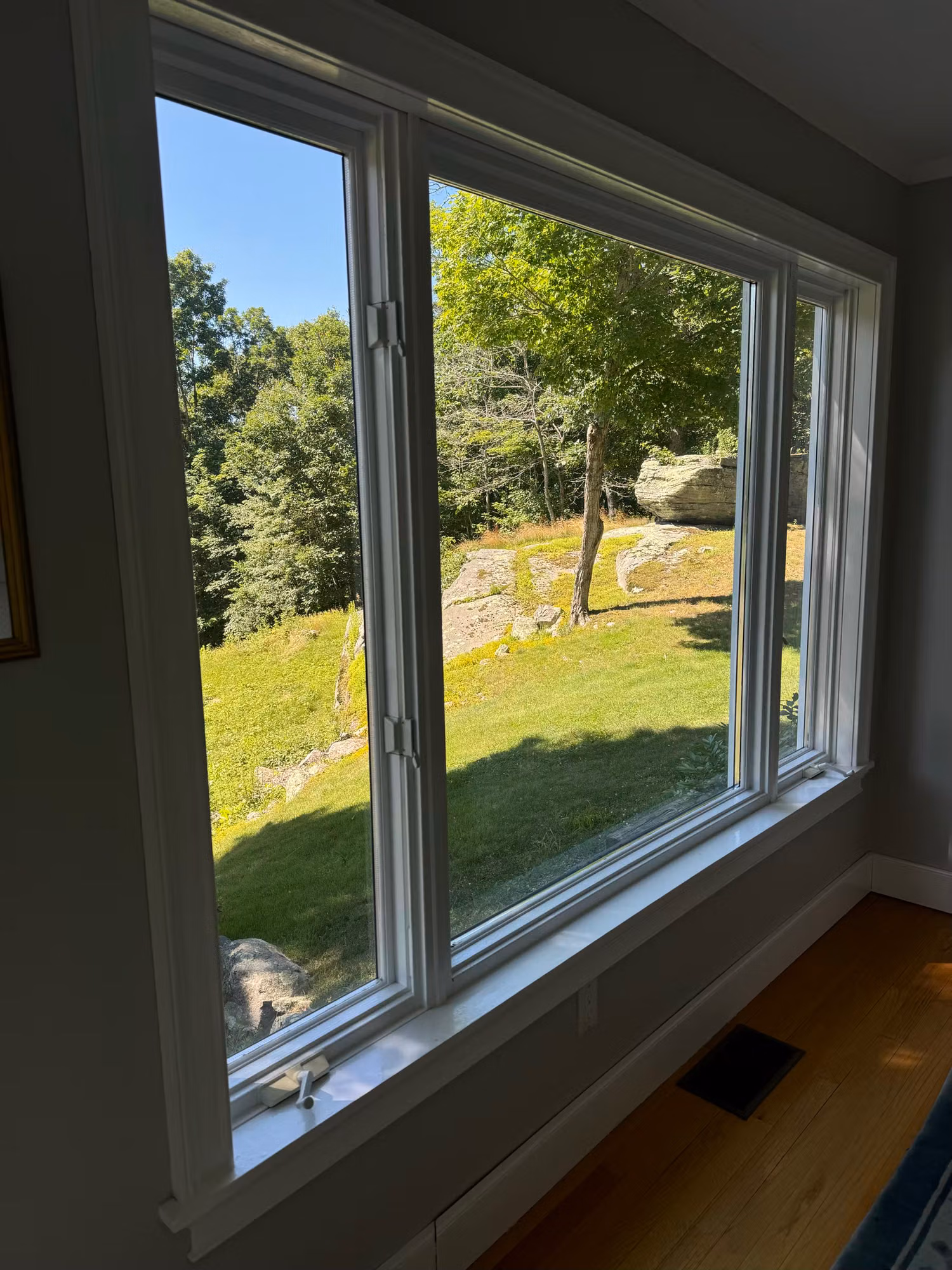 Three-panel casement window with applied film overlooking a rocky lawn from a hardwood-floored room.