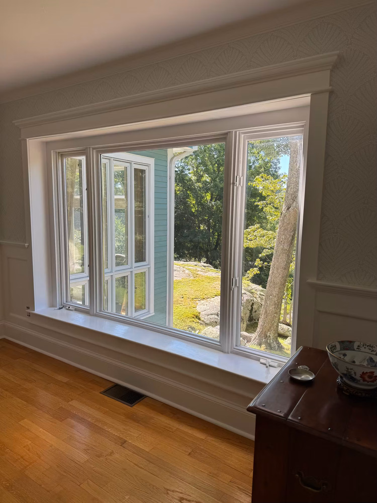 Triple bay window with applied film framed by white trim, overlooking trees beside a wood floor and cabinet.