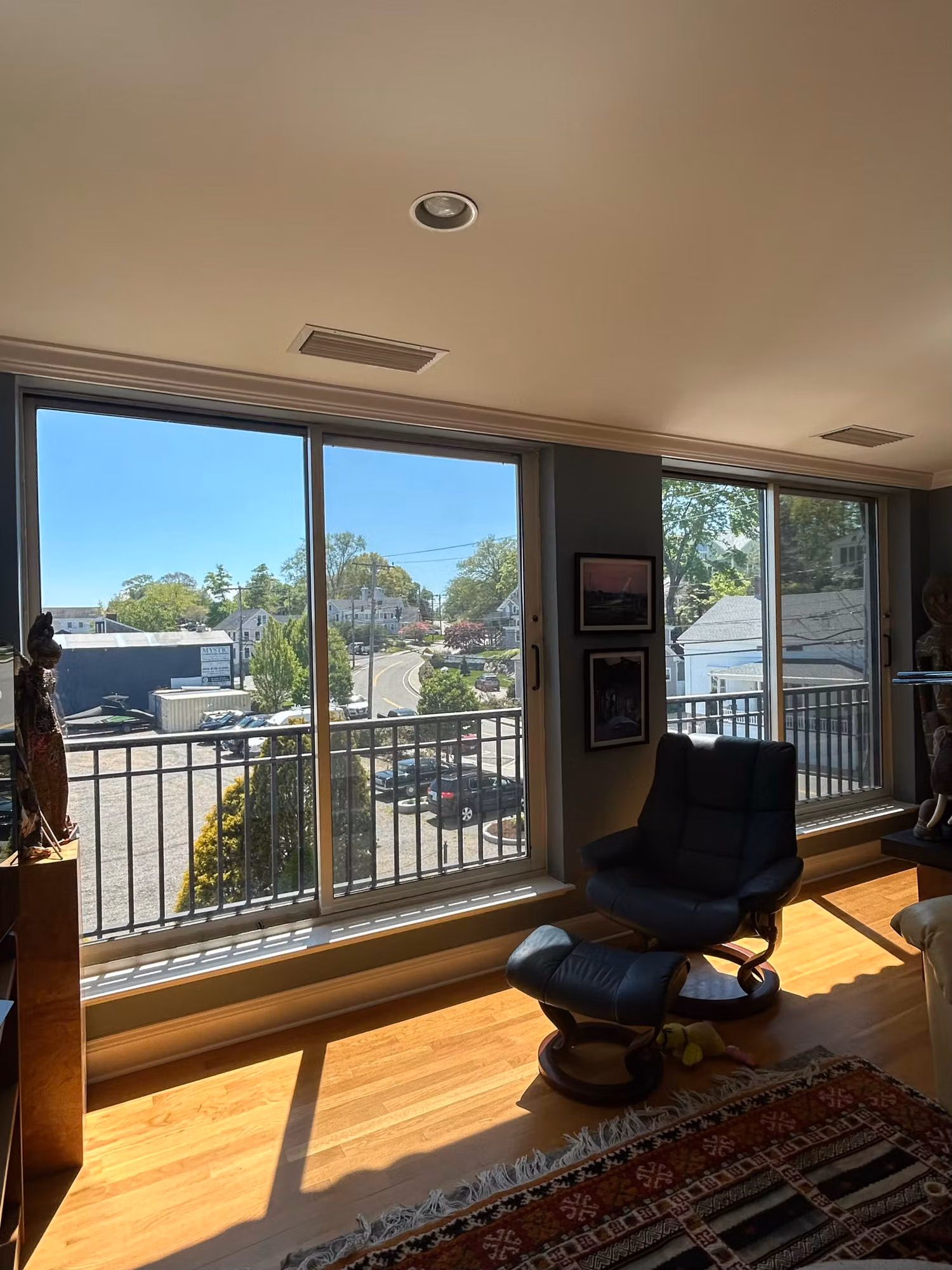 Sunlit living room with sliding glass doors and a balcony recliner, treated glass framing a rooftop town view.