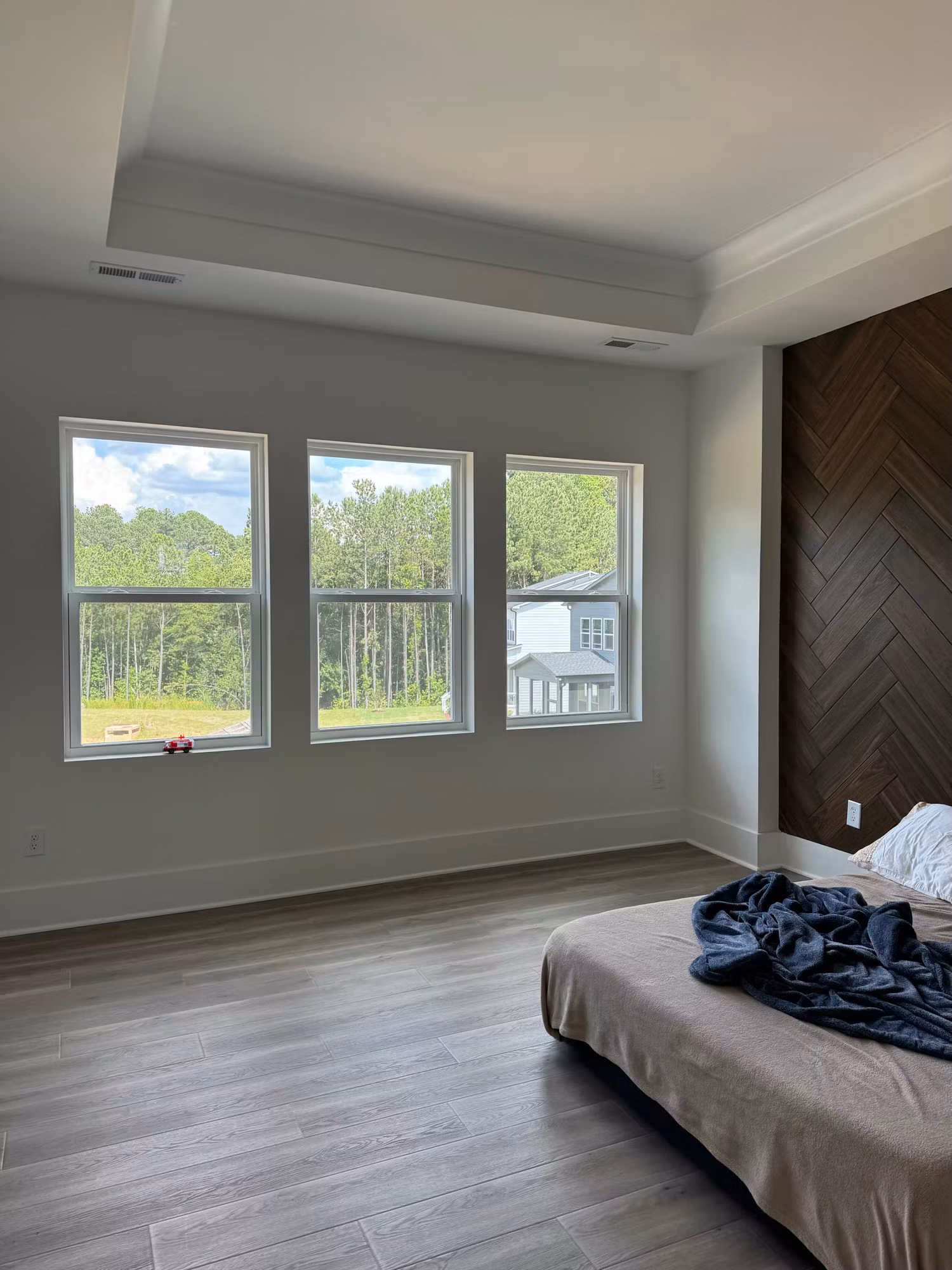 Tray-ceiling bedroom with three tinted picture windows overlooking a wooded backyard and neighboring homes.