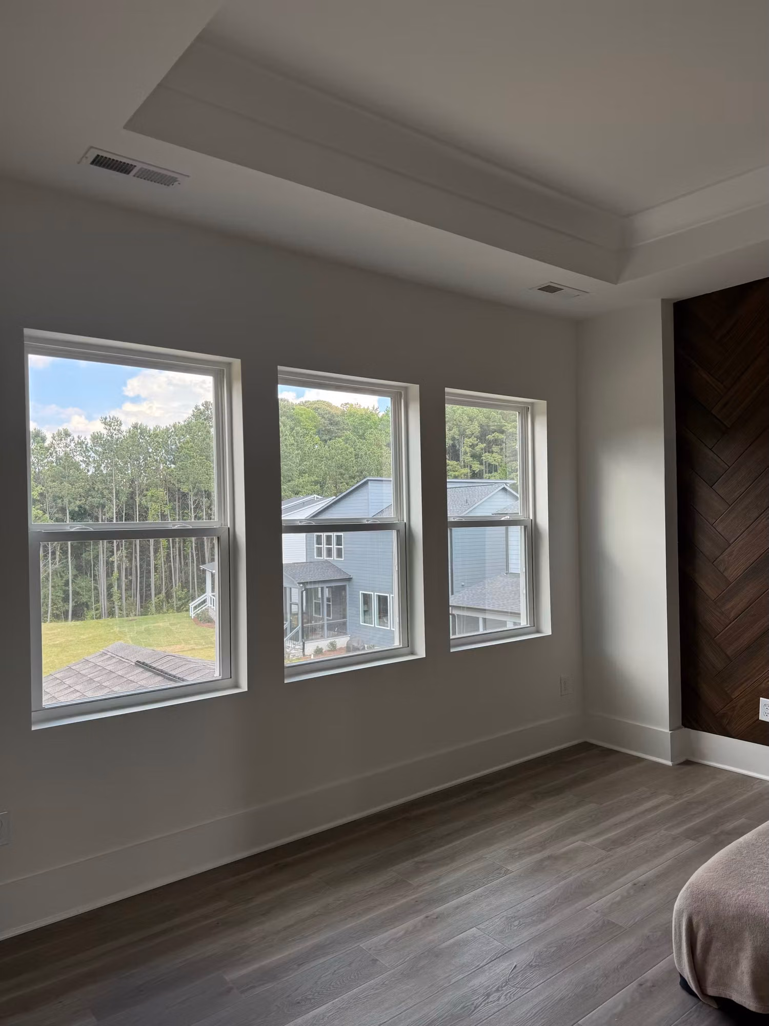 Tray-ceiling bedroom with three tinted windows overlooking neighboring rooftops and a wooded lot.