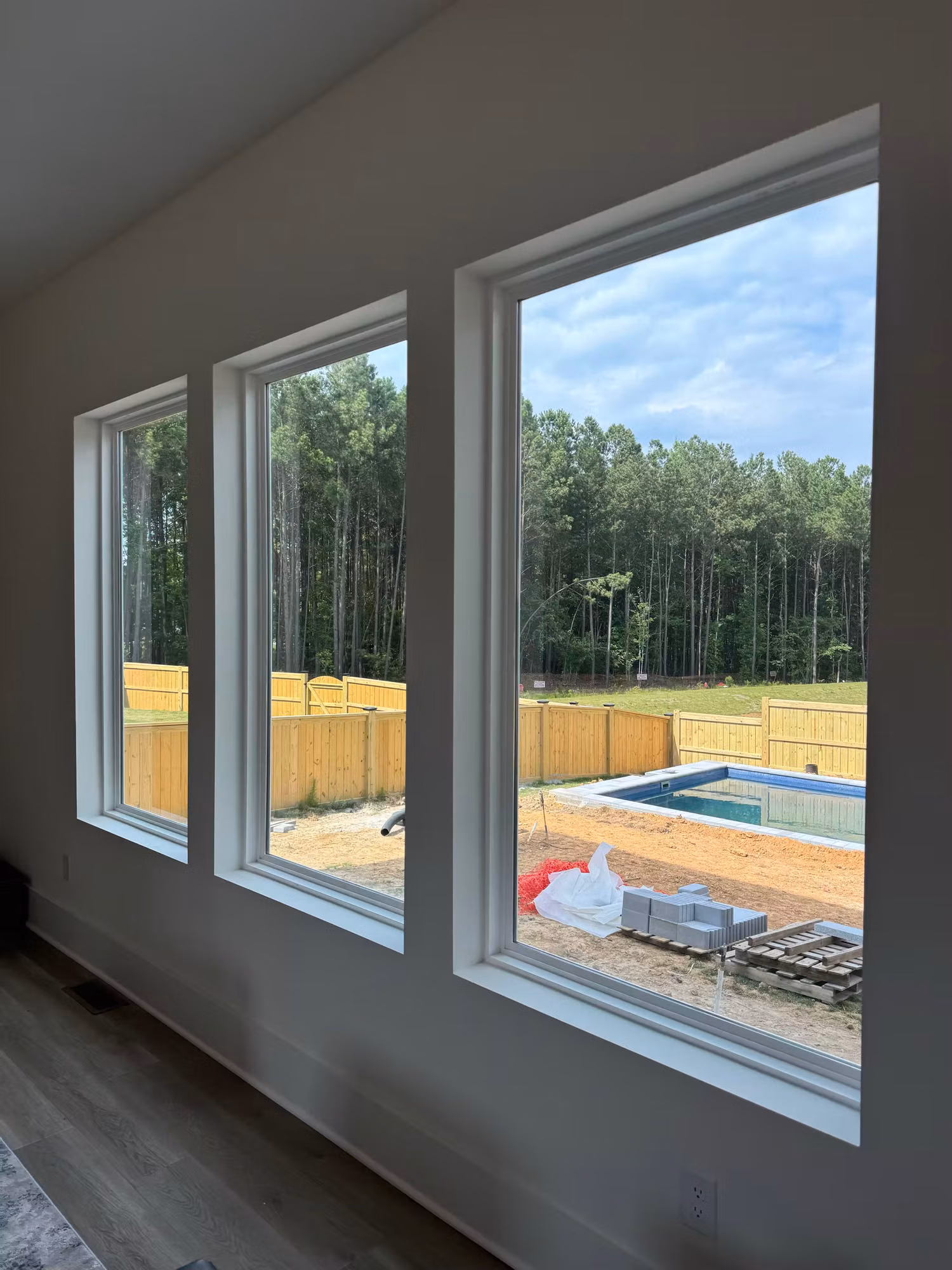 Three large living room windows looking out over a backyard pool and wooded tree line.