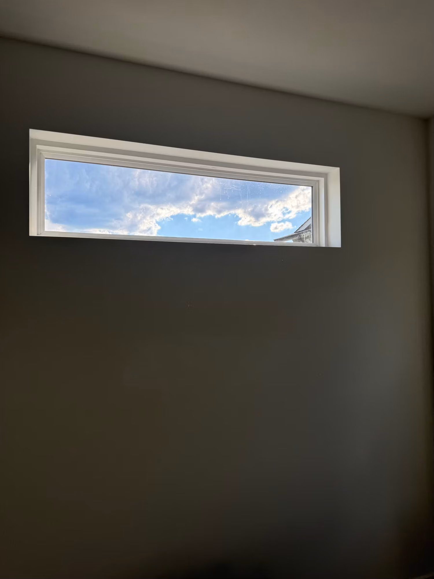 Narrow horizontal transom window set high on a wall showing a cloudy blue sky beyond the glass.