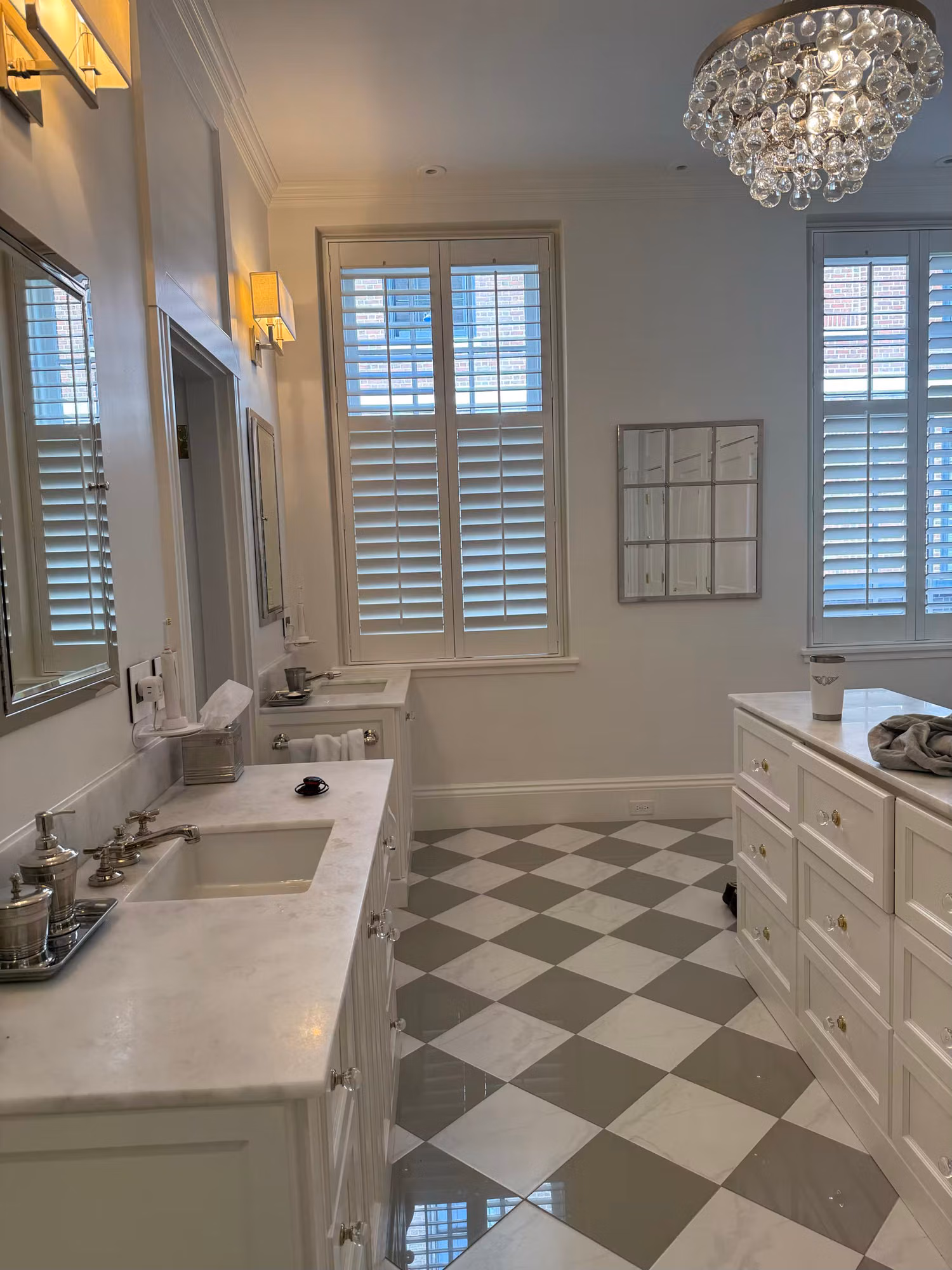 Marble bathroom with clear windows fitted with white plantation shutters and a checkerboard tile floor.