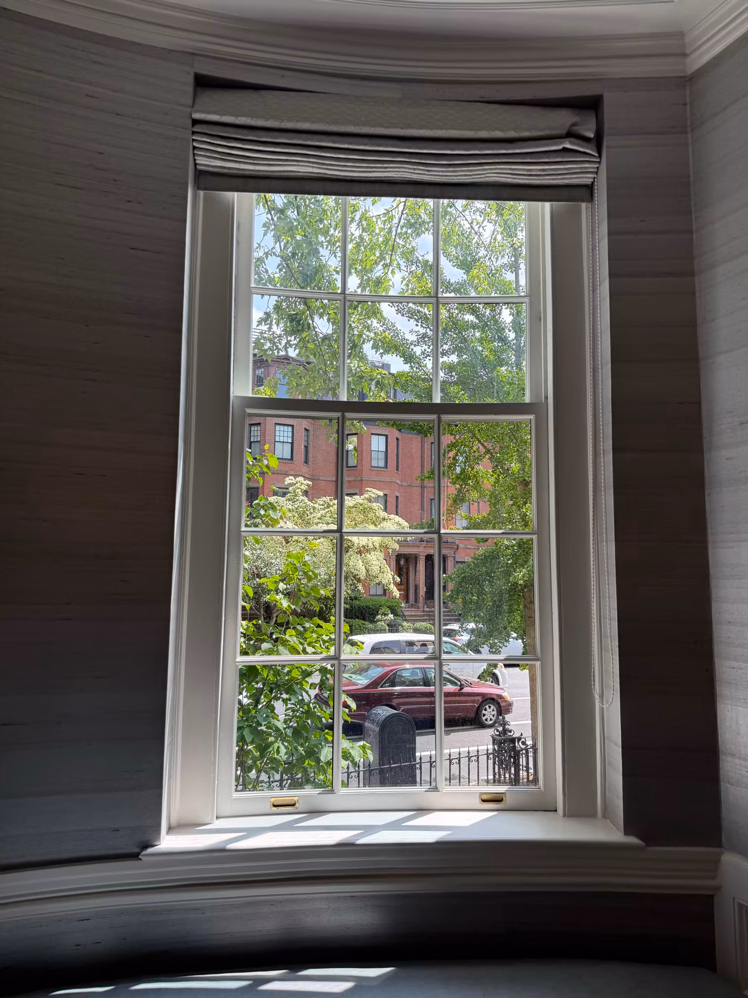 Tall divided-light double-hung window set in a curved grasscloth bay with a street view beyond.