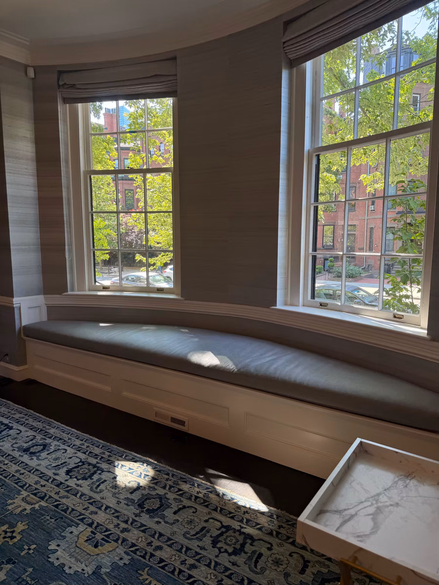 Curved bay with two divided-light windows above an upholstered window seat and grasscloth walls.