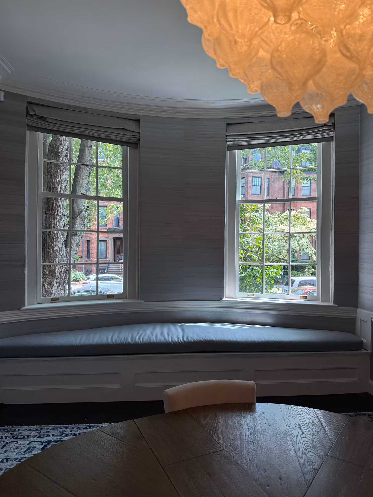Curved dining bay with two divided-light windows above a cushioned window seat and grasscloth walls.