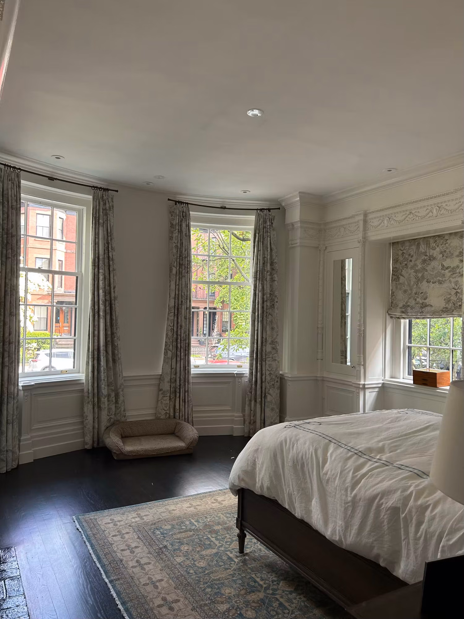 Bedroom with a curved bay of grid windows and floral drapes beside a made bed and an additional shaded window.