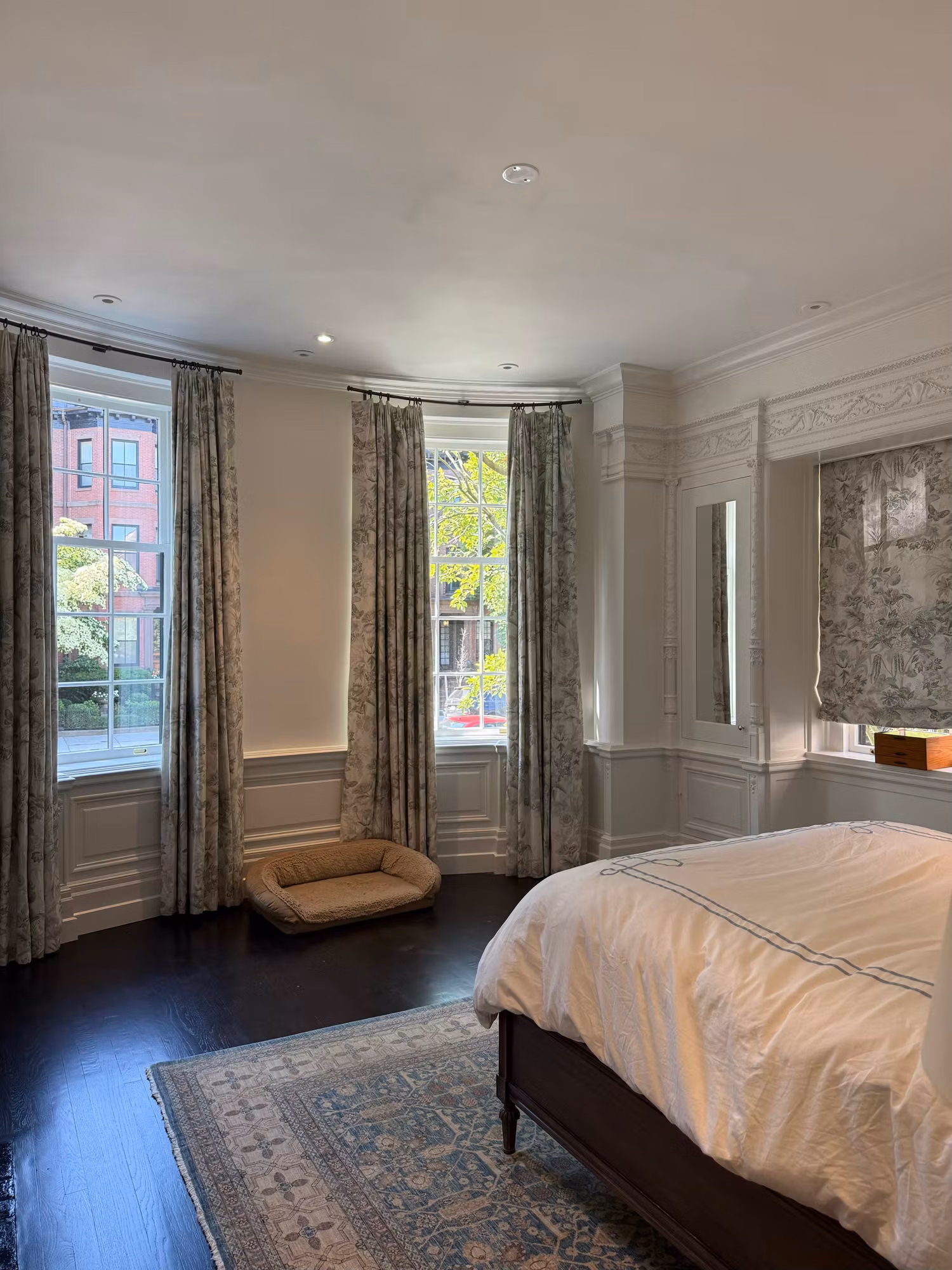 Bedroom bay with clear divided-light sash windows and patterned drapery filtering daylight onto dark wood floors.
