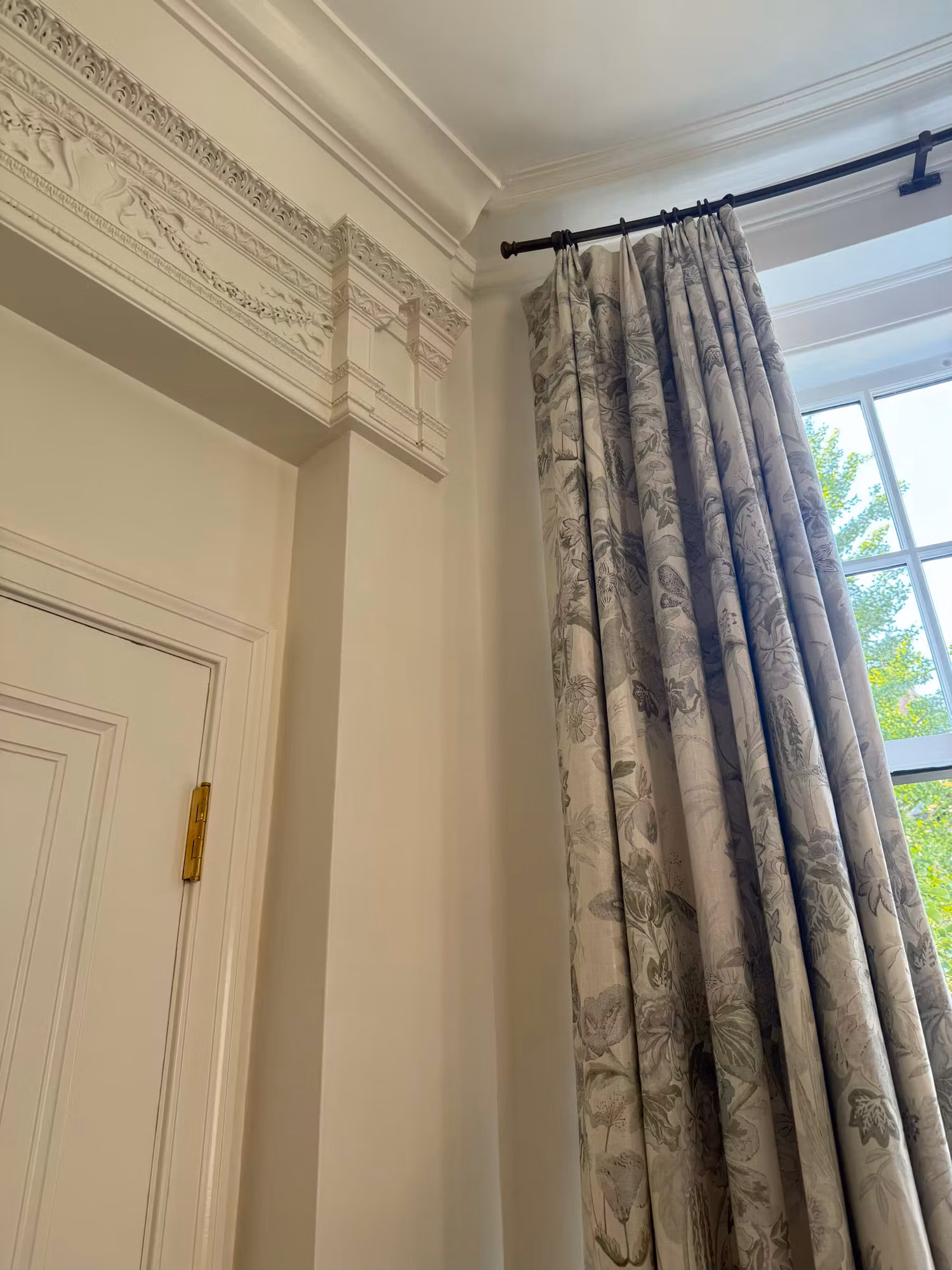 Close view of a tall window edge with carved cornice molding and floral toile drapery beside a paneled door.