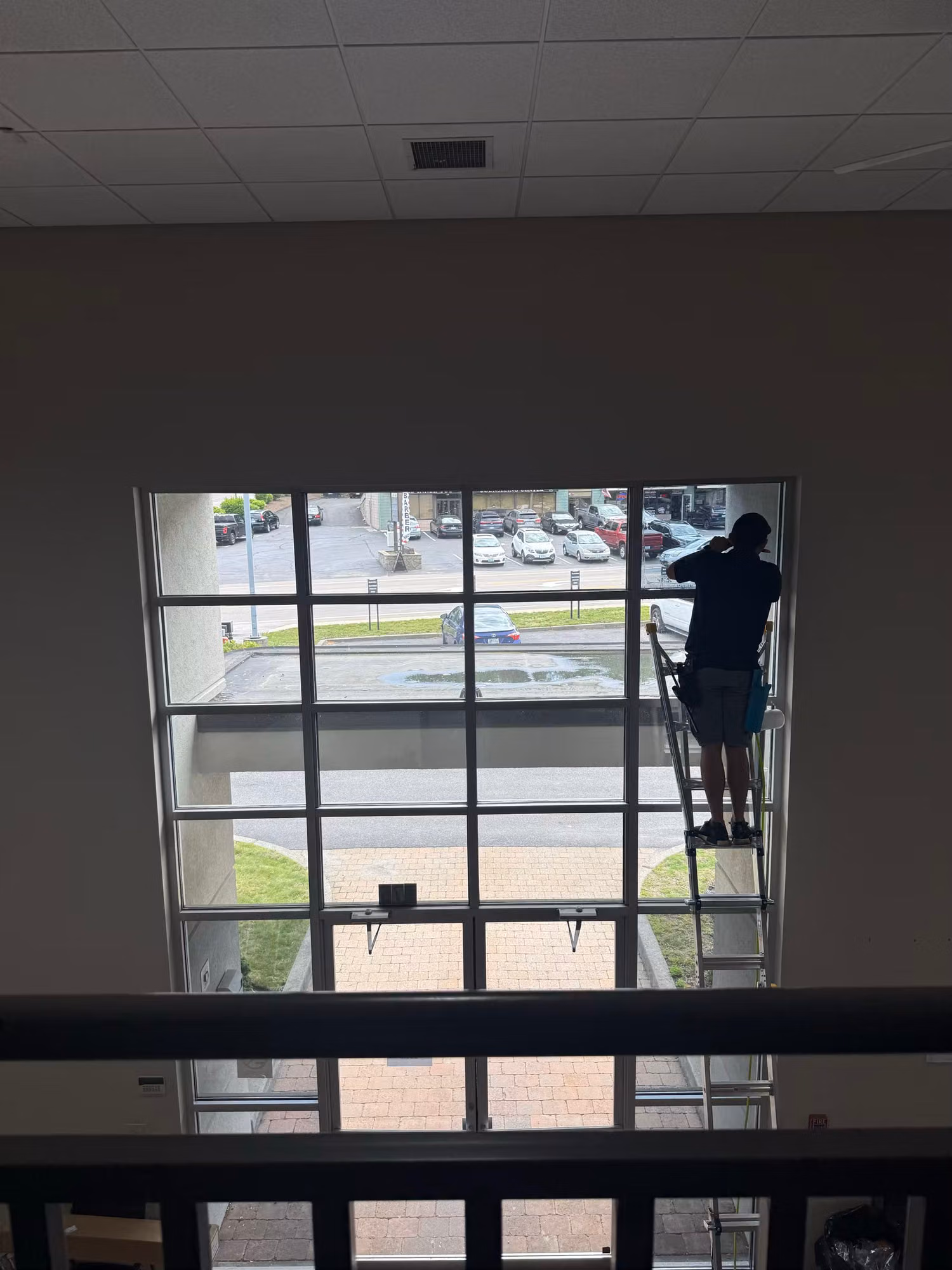 Worker on a ladder installs window film on a large gridded curtain-wall window overlooking a parking lot from an interior landing.