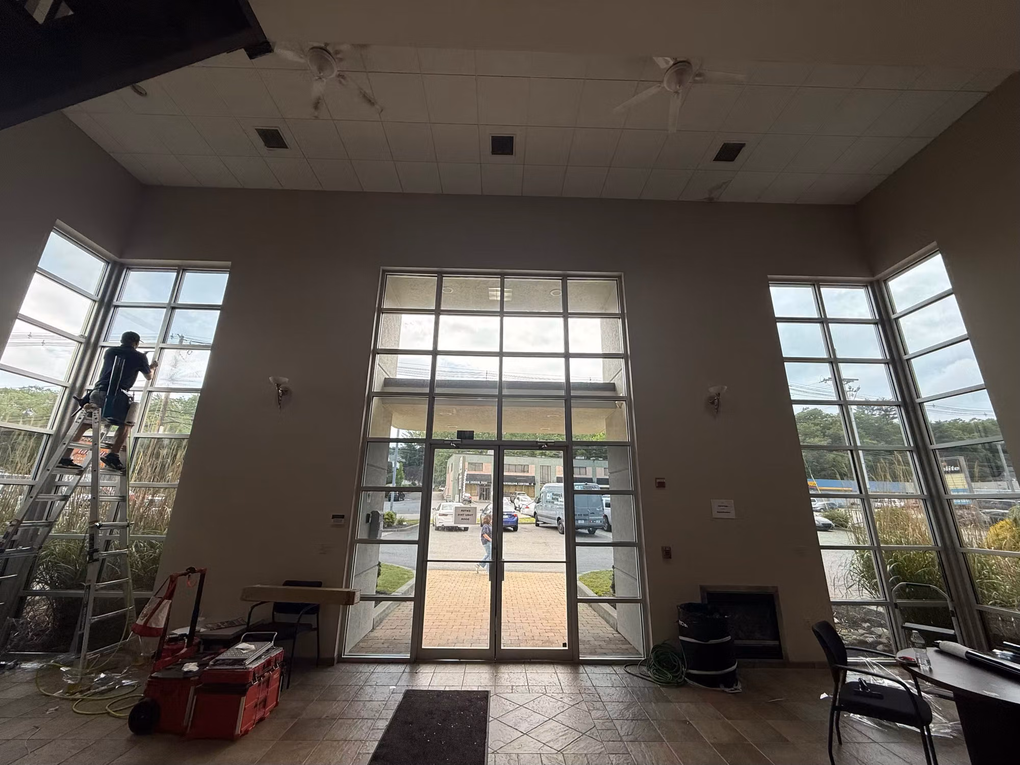 Installer on a ladder applies film to a tall glass curtain-wall window beside double glass entry doors in a commercial lobby.