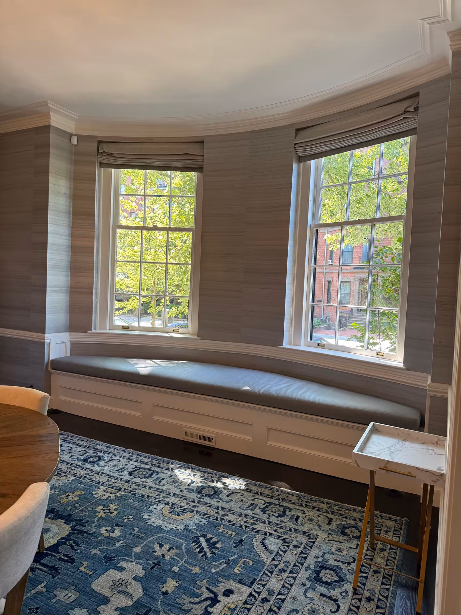 Curved dining nook bay with clear divided-light windows above an upholstered window seat and grasscloth walls.