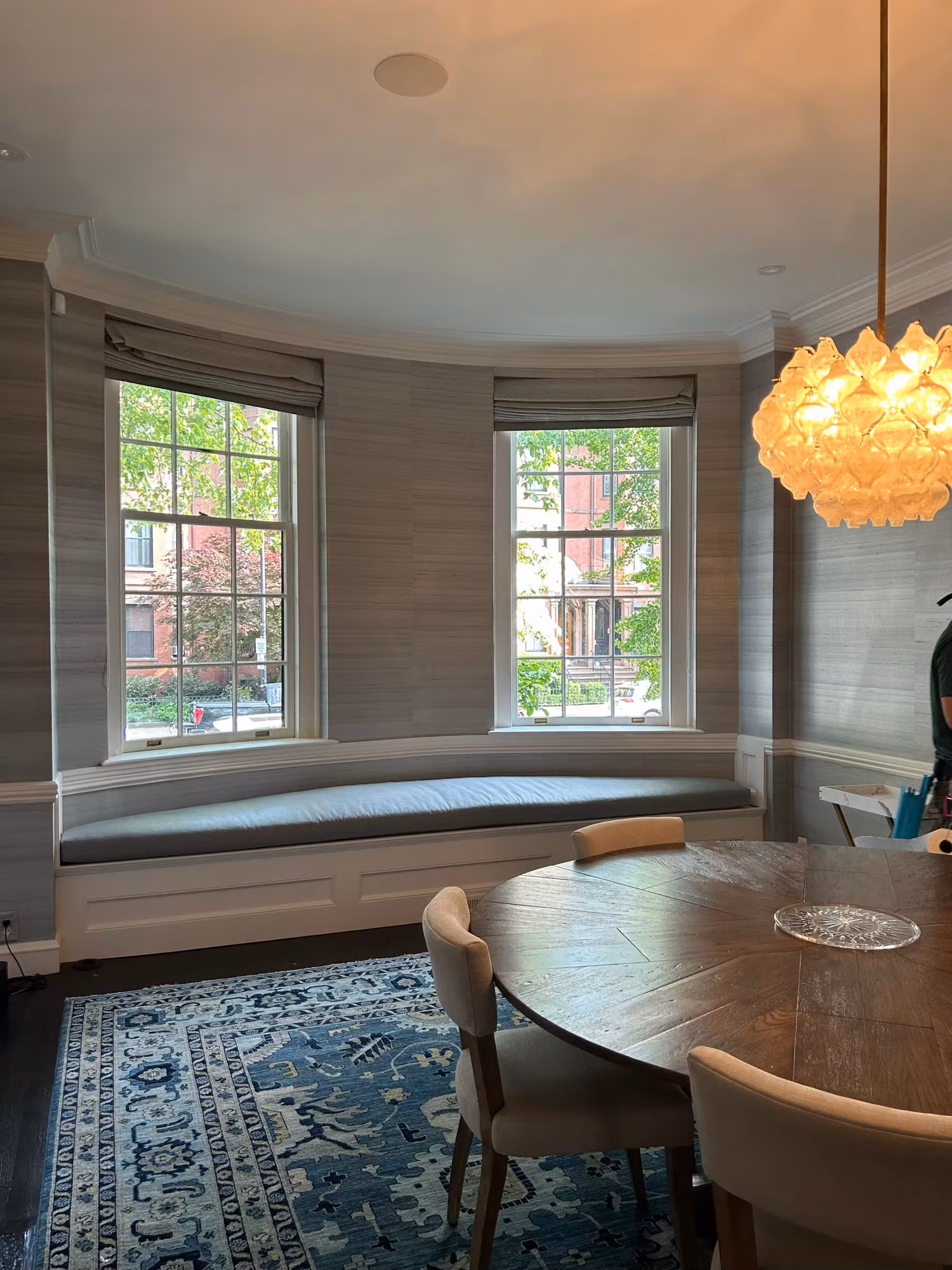 Dining room curved bay with two grid windows over a cushioned window seat, roman shades, and a round wood table.